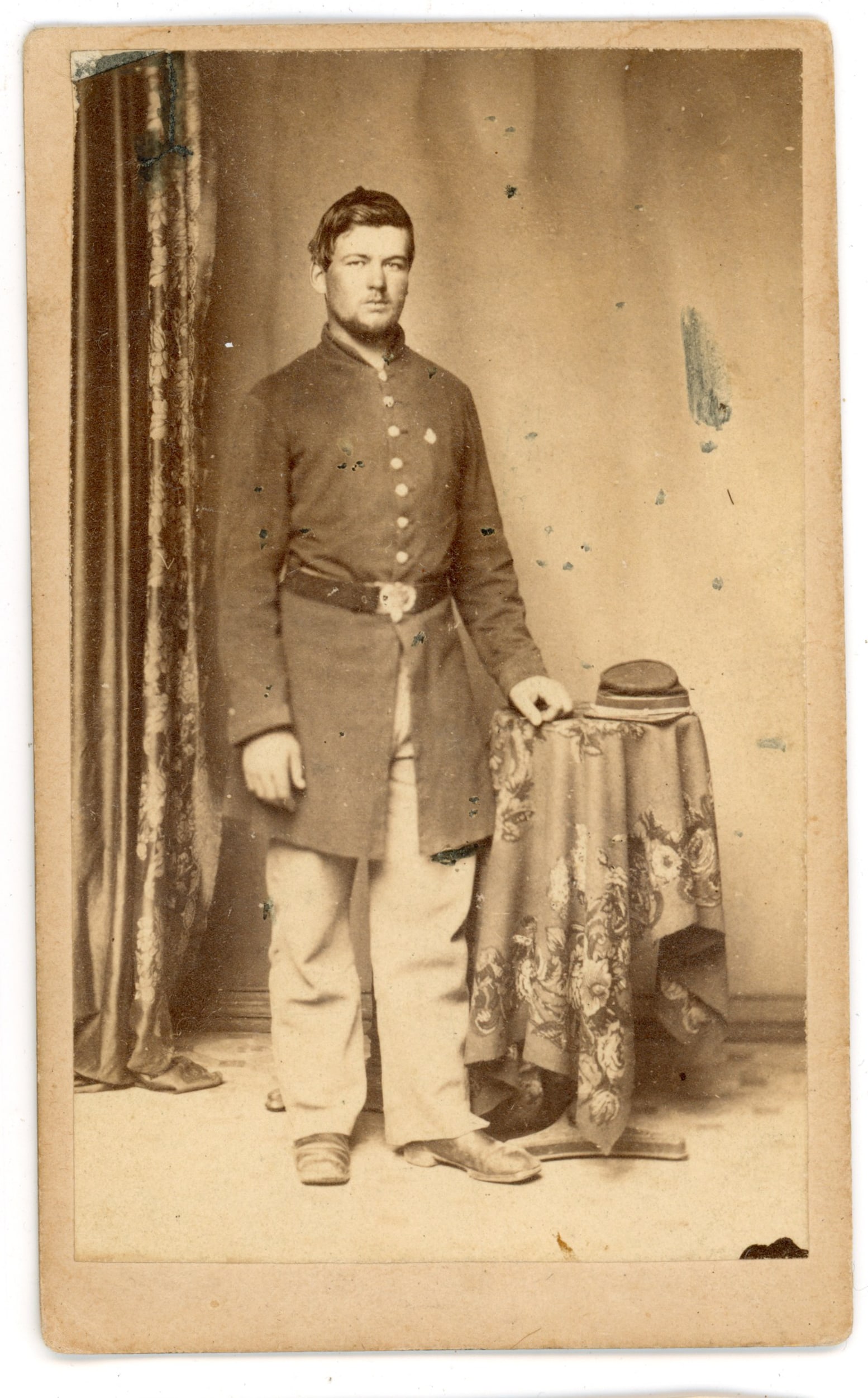 CDV Union Soldier with Kepi Wolff Gallery Alexandria: Carte de visite portrait of an unidentified Union soldier wearing a kepi and frock coat, standing beside a small draped table with his hat resting on the tabletop. The soldier’s belt plate and unifo