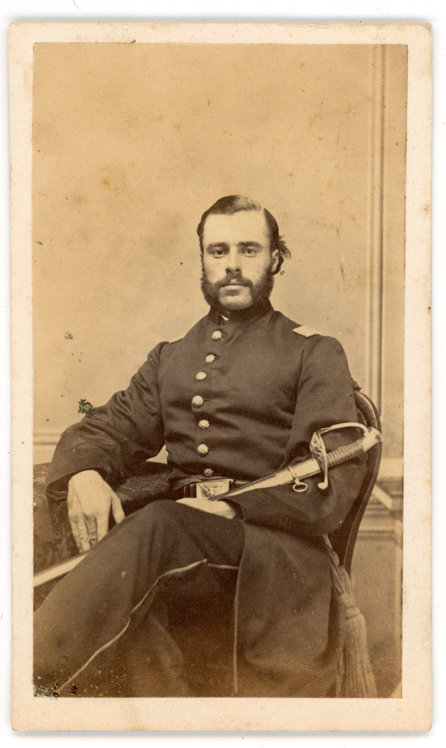 CDV Union Officer Seated Portrait: Carte de visite portrait depicting an unidentified Union Army officer seated in studio wearing a dark officer’s frock coat with shoulder straps and sword belt. The officer rests one arm on the chair