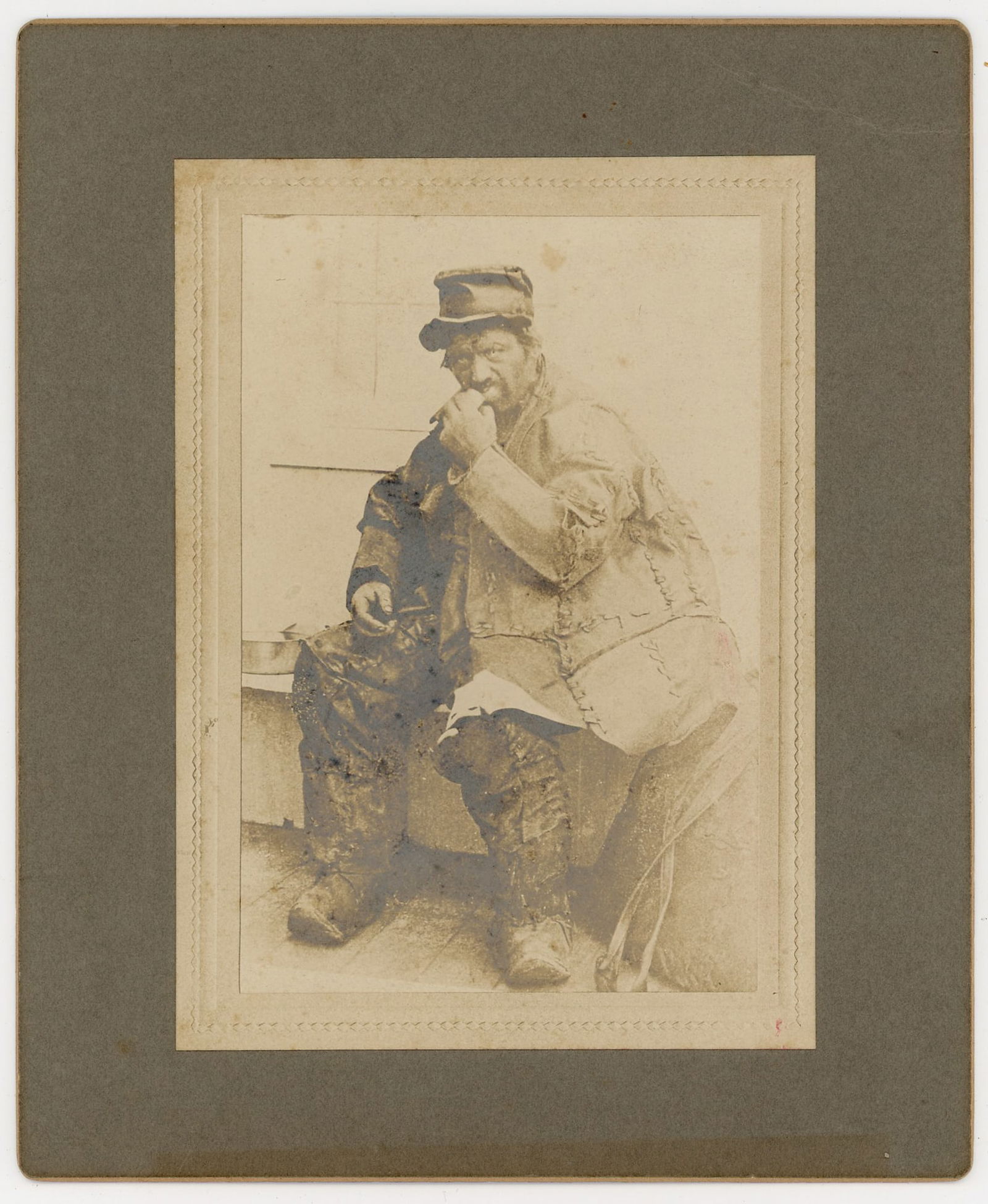 Mounted Photograph of "Leatherman"Smoking Pipe: Mounted albumen photograph depicting famous New England vagabond "Leatherman" seated wearing a cap and heavy patched clothing, shown smoking a pipe while resting on a wooden bench. The image appears t
