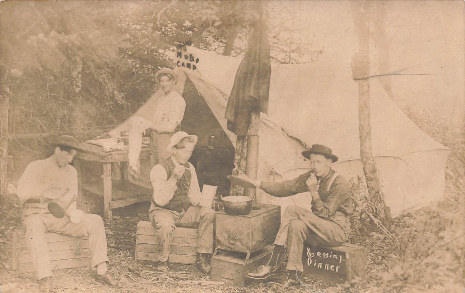 RPPC Hobo Camp Eating Dinner Postcard: Real photo postcard showing a group of men at an outdoor camp, with a prominent sign reading “HOBO CAMP” and “EATING DINNER.” The scene is arranged around a makeshift cooking area with