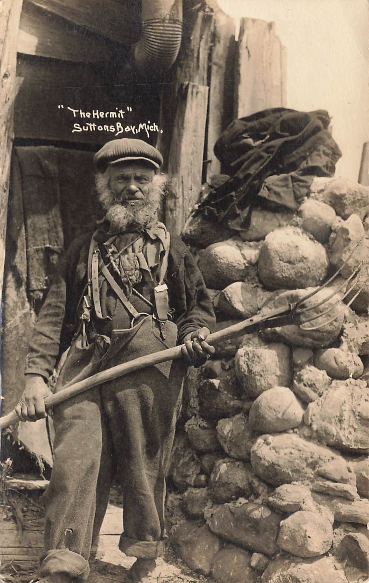 RPPC “The Hermit?? Suttons Bay, Mich.: Real photo postcard labeled “The Hermit” at Suttons Bay, Michigan, showing a bearded man posed in front of a rustic cabin. The man stands holding a long-handled tool, dressed in work clothes a