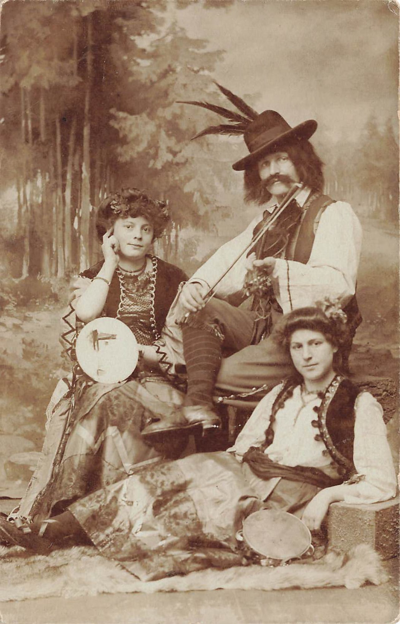 RPPC Costumed Trio With Violin Postcard: Real photo postcard showing three costumed performers posed in a studio setting, including a man holding a violin and two women holding tambourines. The man wears a feathered hat and cape over a light