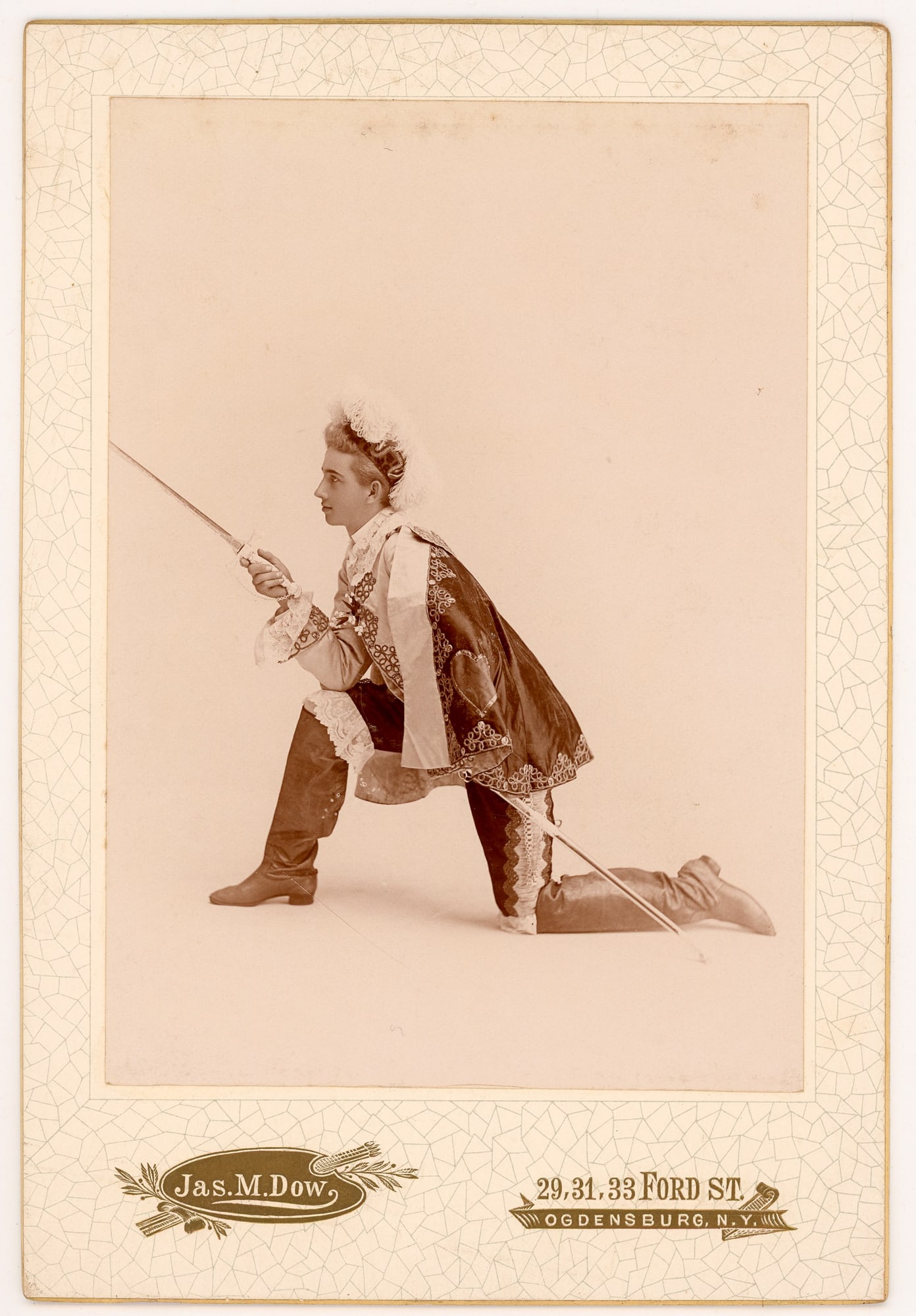 Jas. M. Dow Cabinet Card Costumed Youth with Sword: Cabinet portrait of a young male posed in theatrical costume, kneeling on one knee and holding a drawn sword. The subject wears a feathered cap, embroidered doublet with decorative braid, lace cuffs,