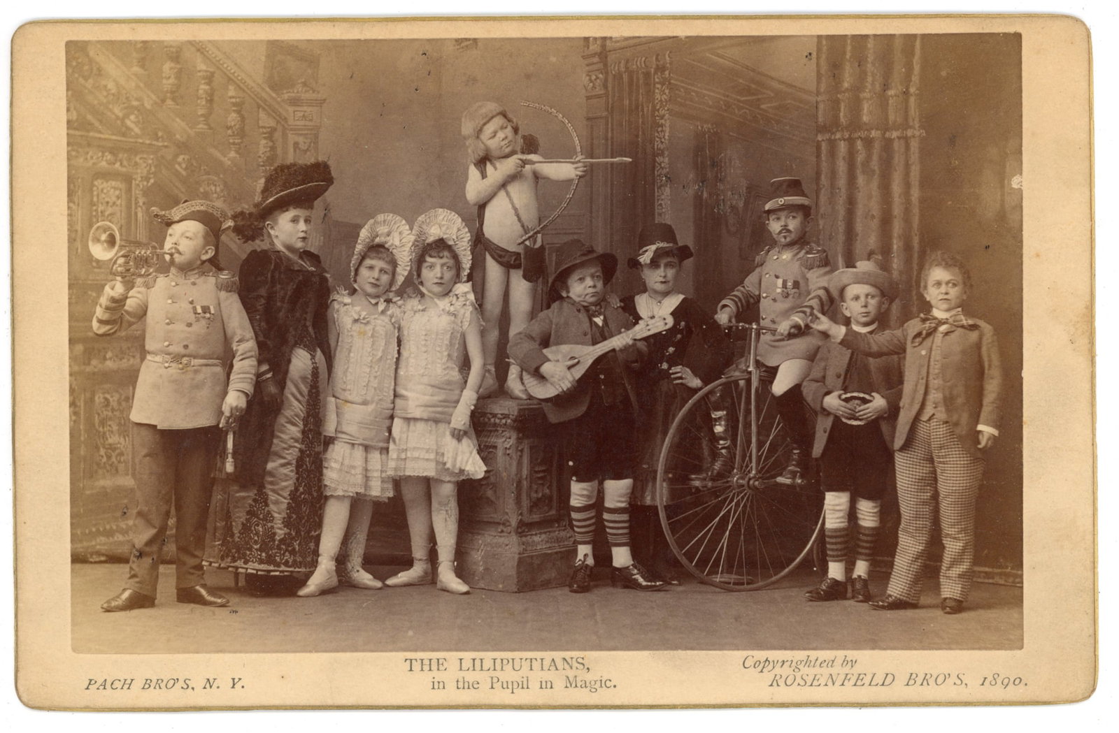 Pach Bros Cabinet Card Lilliputians Pupil in Magic Horn: Cabinet card group portrait titled “The Lilliputians, in the Pupil in Magic.” The photograph shows a troupe of little person performers posed in a theatrical studio interior with architectural