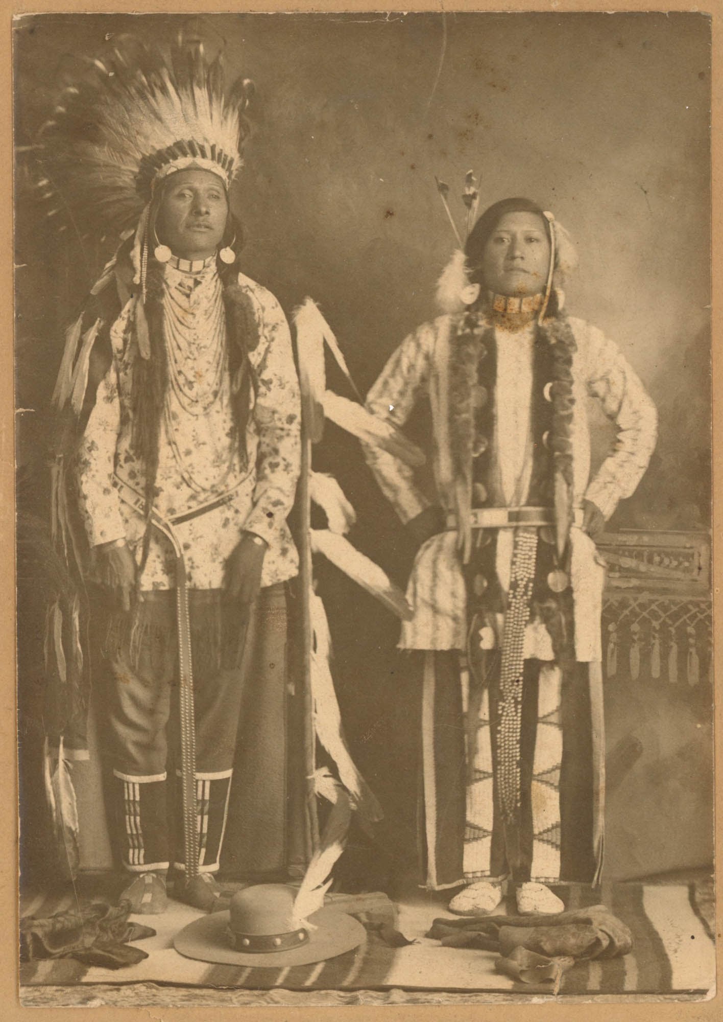 Cabinet Card, Two Unidentified Native American Men, Full Length: Two unidentified Native American men stand full length before a plain studio backdrop in this albumen photograph mounted on a dark green cabinet card. The man at left wears a full eagle feather headdr
