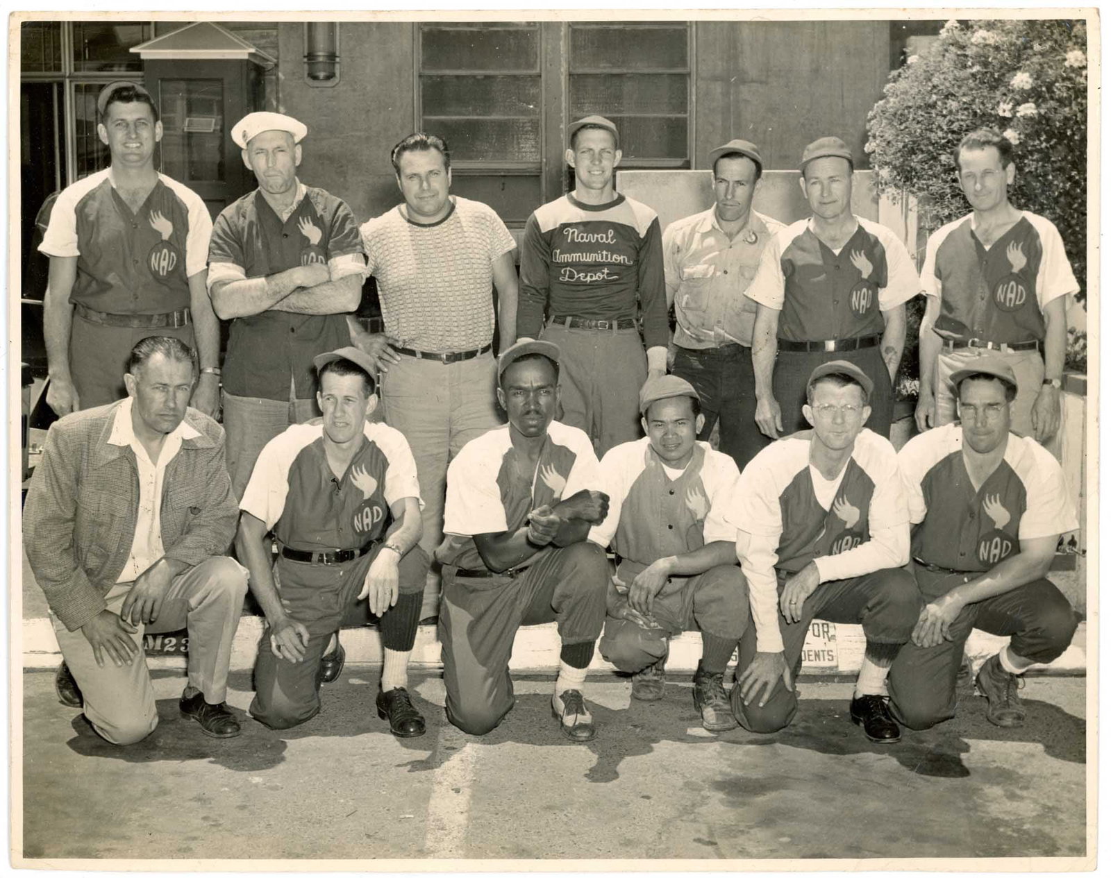 Photograph, Integrated Baseball Team, African American, Nebraska: Thirteen members of the Naval Ammunition Depot intra-base baseball team pose in two rows outside a depot building in this gelatin silver print, identified on the reverse as the NAD team at Hastings, N