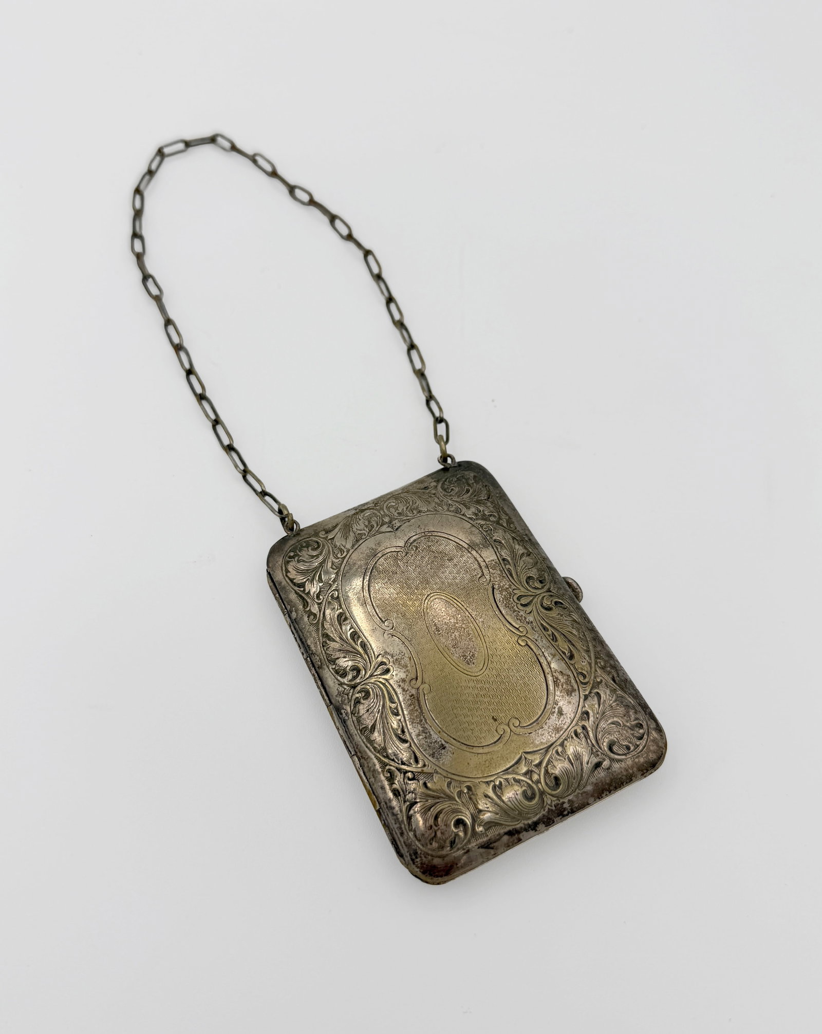Silverplated Metal Purse: Early 20th-century dance purse vanity compact in engraved silver-tone metal, featuring floral and foliate scrollwork surrounding an unmonogrammed oval cartouche. The hinged case has a chain handle