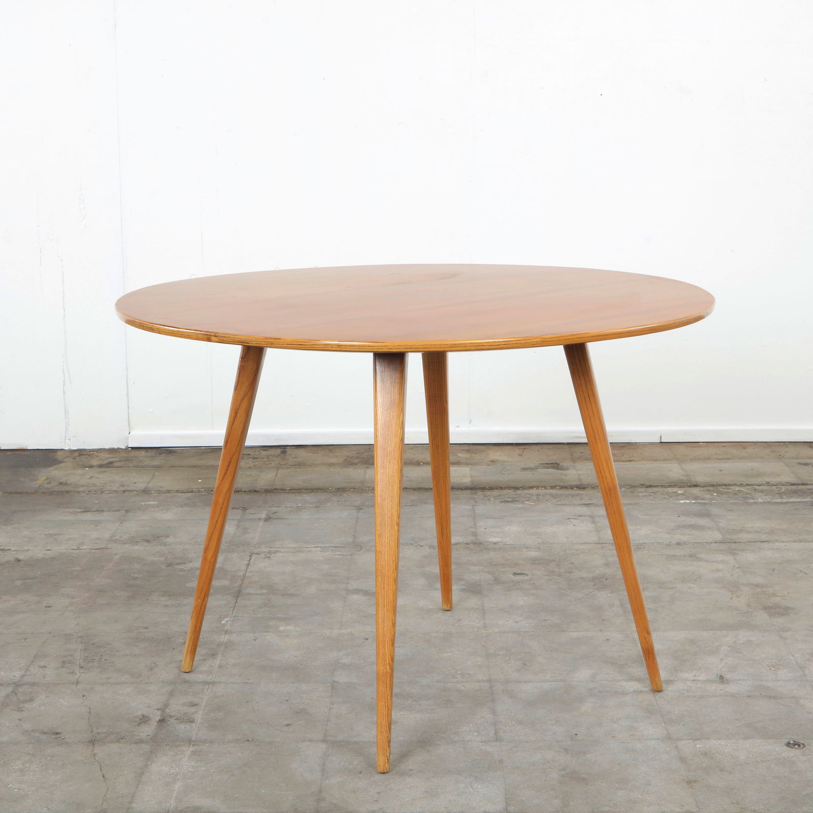 Mid-Century Wood Dining Table in the style of Ercol, 1960s: Round dining table in natural wood, raised on tapered legs. Its minimalist form recalls the enduring mid-century aesthetic. Height: 30 in. Diameter: 43.5 in.