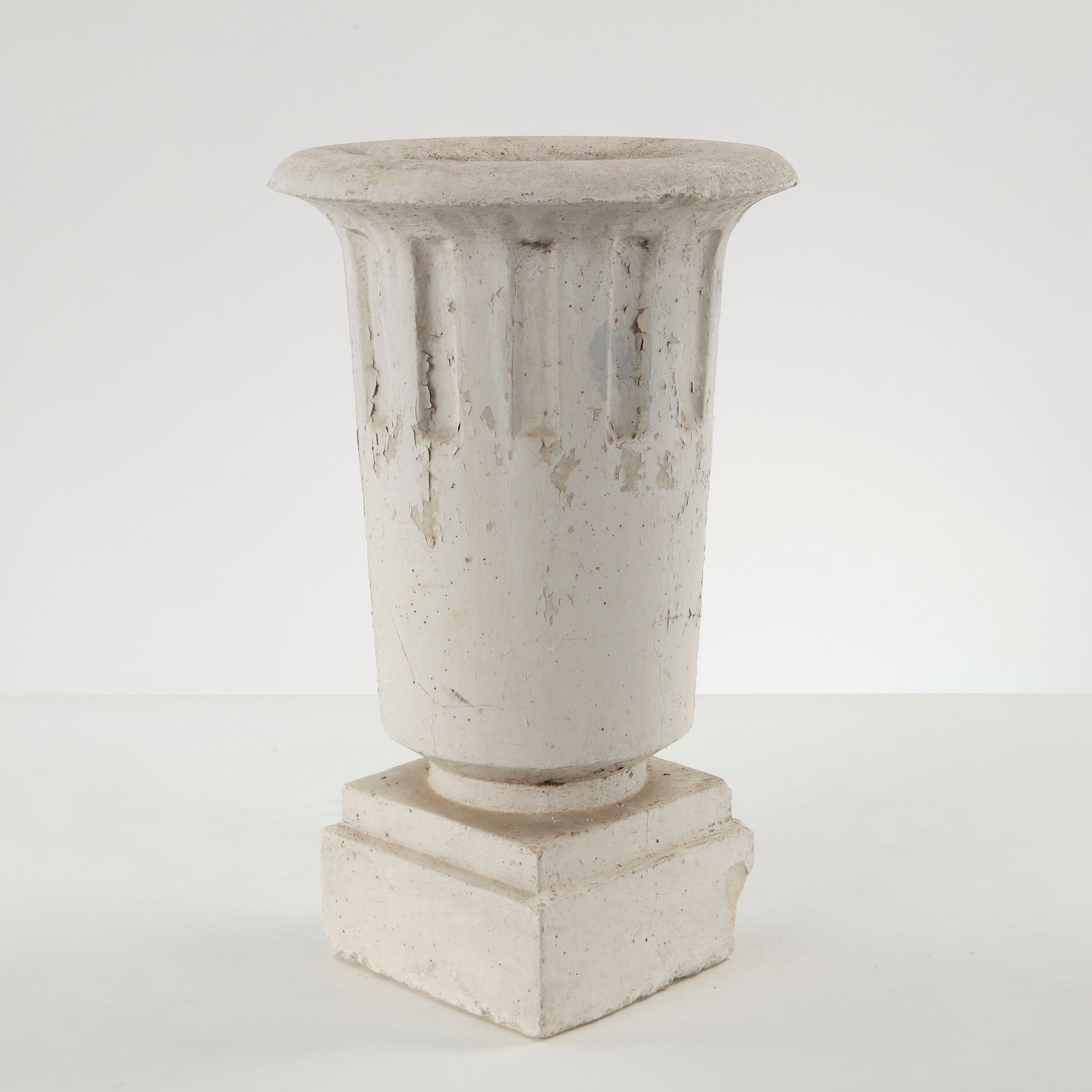Fluted Pedestal Urn, 1950s: Cast stone urn with fluted body raised on a stepped square pedestal. The restrained classical profile is animated by vertical fluting and softened by traces of time and use. A timeless garden ornament