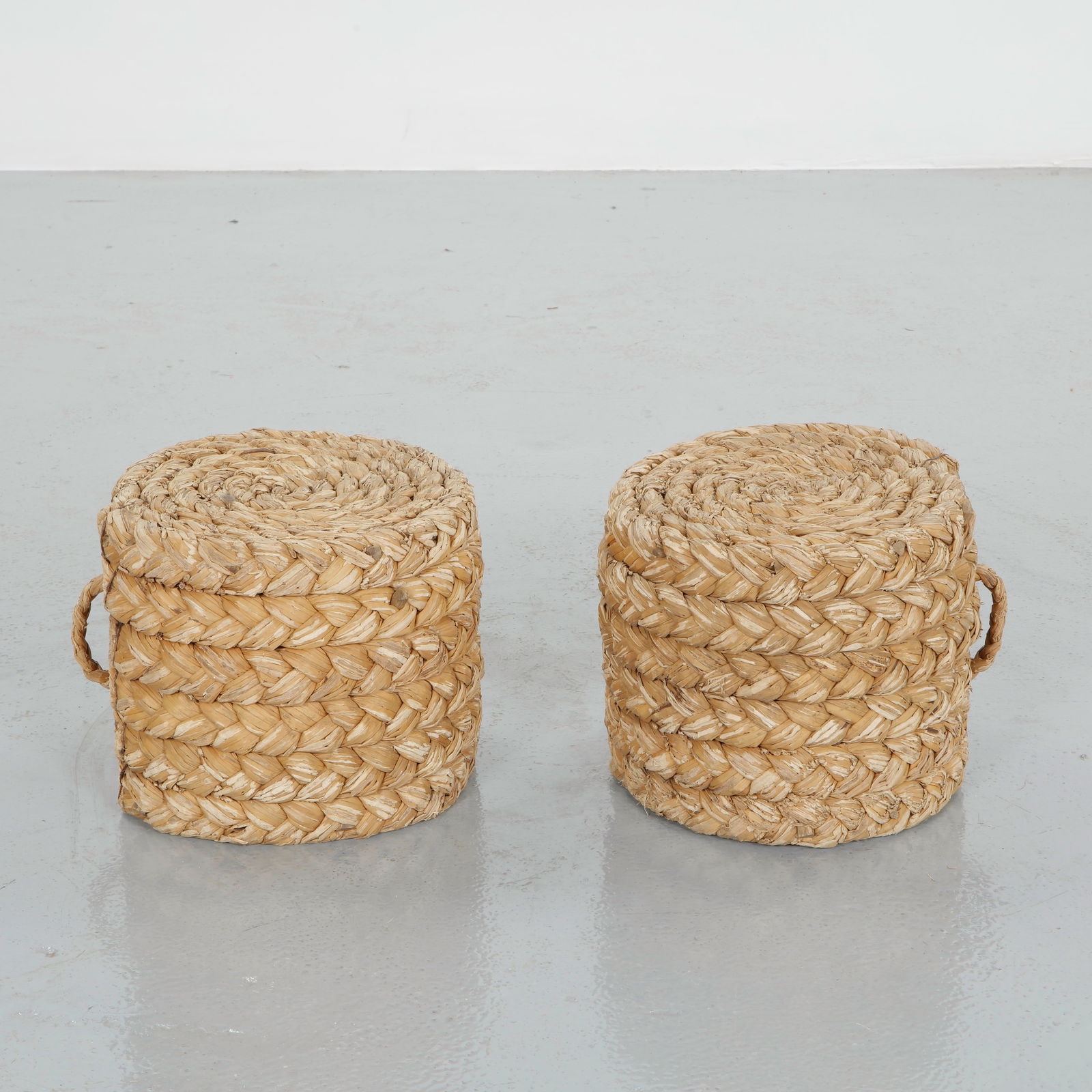 Pair of Rope Stools in the style of Audoux Minet, 1950s: Pair of cylindrical stools hand woven in thick braided rope, tightly coiled in concentric spirals to form both seat and body. The robust vegetal fibers and integrated side handles reflect the rustic m