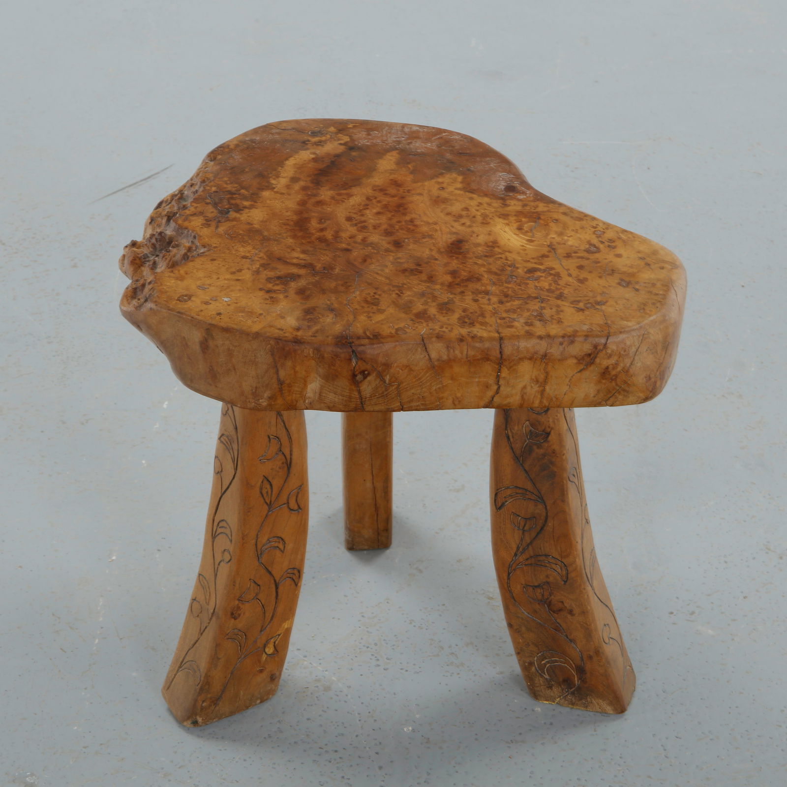 French Brutalist, Carved Burl Wood Low Stool, 1950s: Carved from a thick burl section with free-form contour, this low stool rests on three sculpted legs delicately incised with foliate motifs. The richly figured surface and hand worked details emphasiz