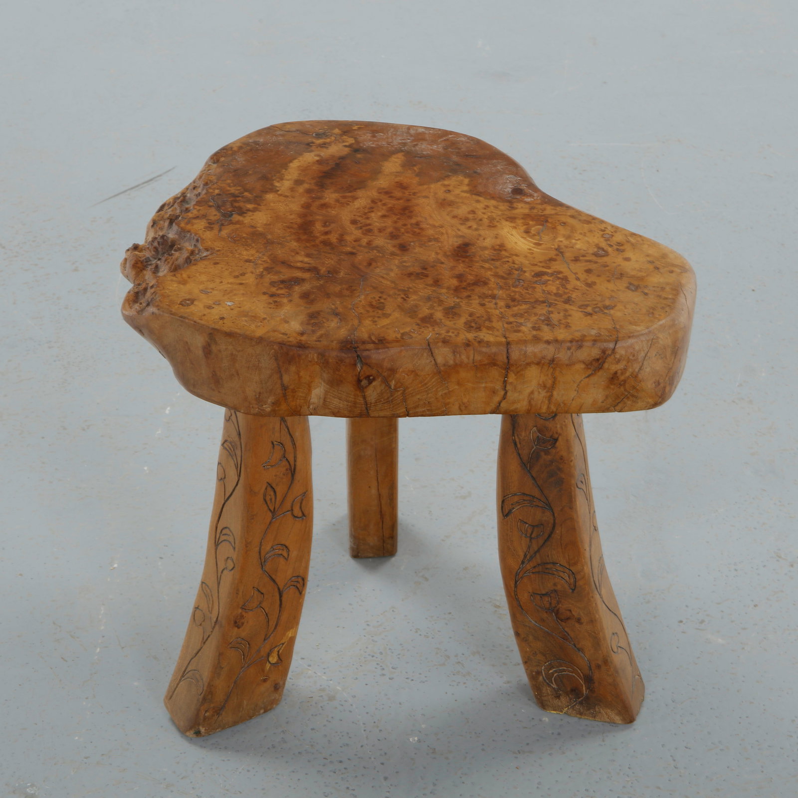French Brutalist, Carved Burl Wood Low Stool, 1950s: Carved from a thick burl section with free-form contour, this low stool rests on three sculpted legs delicately incised with foliate motifs. The richly figured surface and hand worked details emphasiz