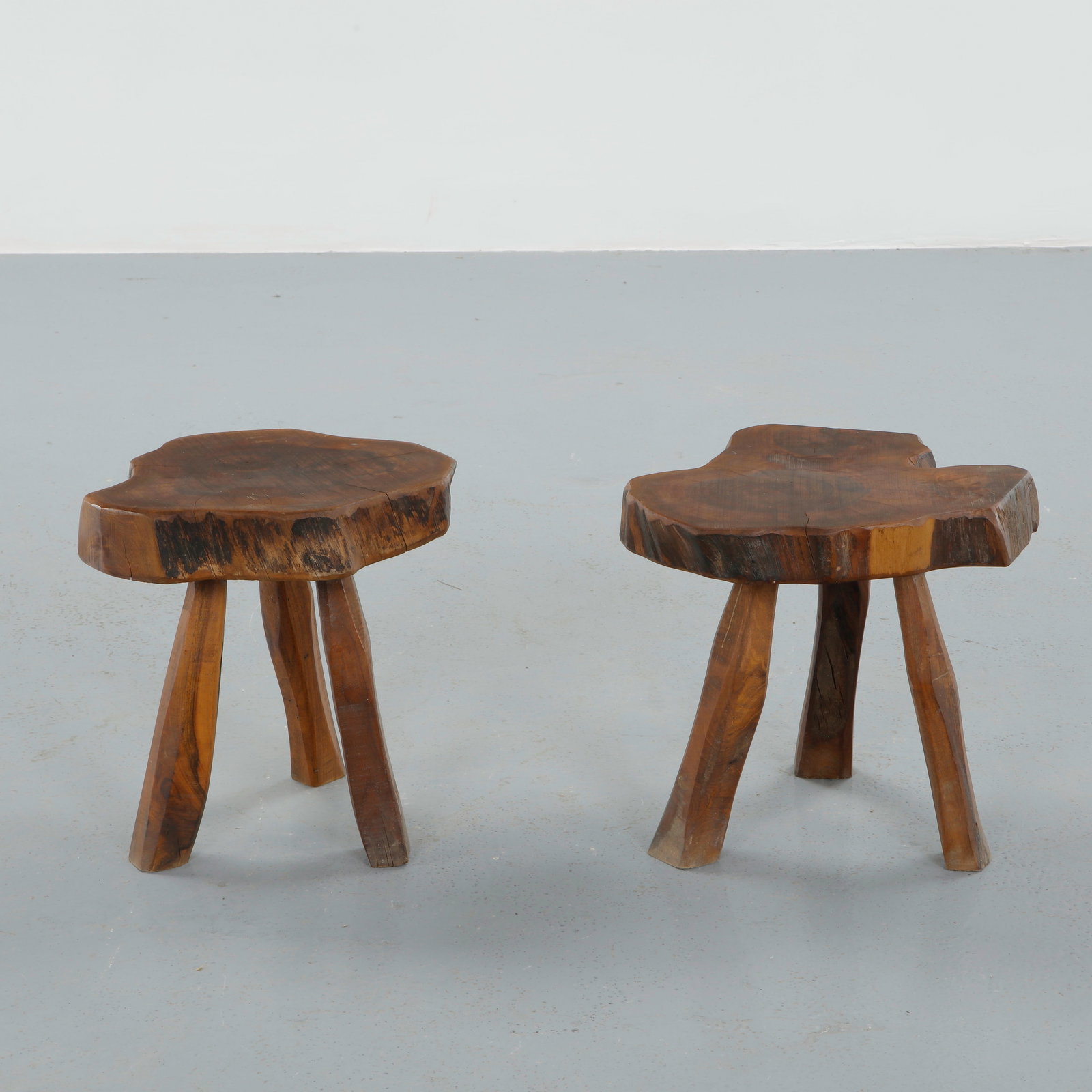 Pair of Brutalist Wood Stools, 1950s: Each carved from a solid section of timber, these stools feature thick organic seats with natural live edges resting on three hand-shaped legs. The expressive grain and irregular contours celebrate ra