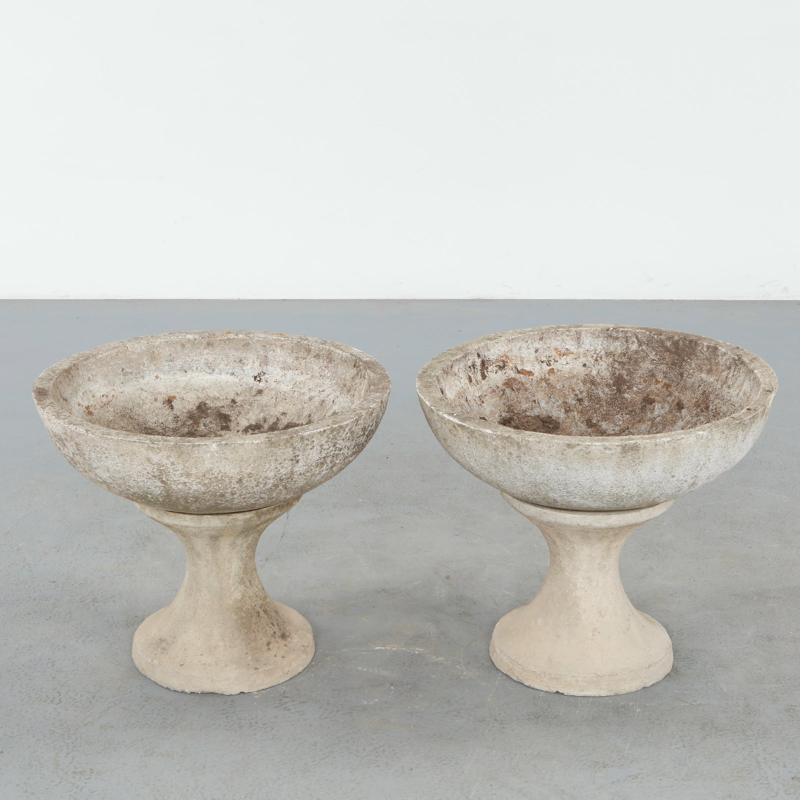 Pair of Stone Planters on Stands, 1960s: Pair of stone bowl planters resting on sculptural pedestal stands with softly curved profiles. The minimalist forms and aged surfaces exemplify the understated elegance of midcentury garden design. He