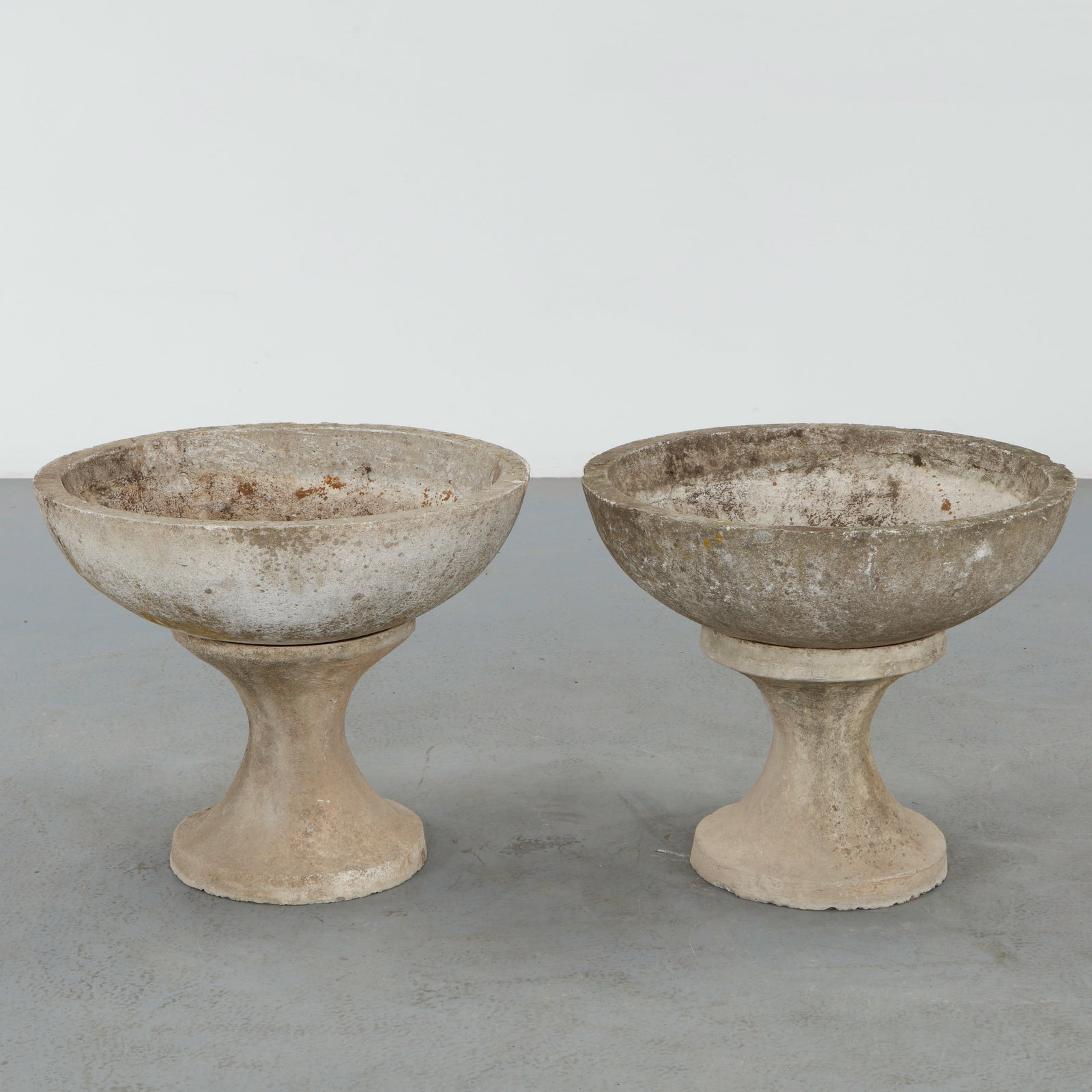 Pair of Stone Planters on Stands, 1960s: Pair of stone bowl planters resting on sculptural pedestal stands with softly curved profiles. The minimalist forms and aged surfaces exemplify the understated elegance of midcentury garden design. He