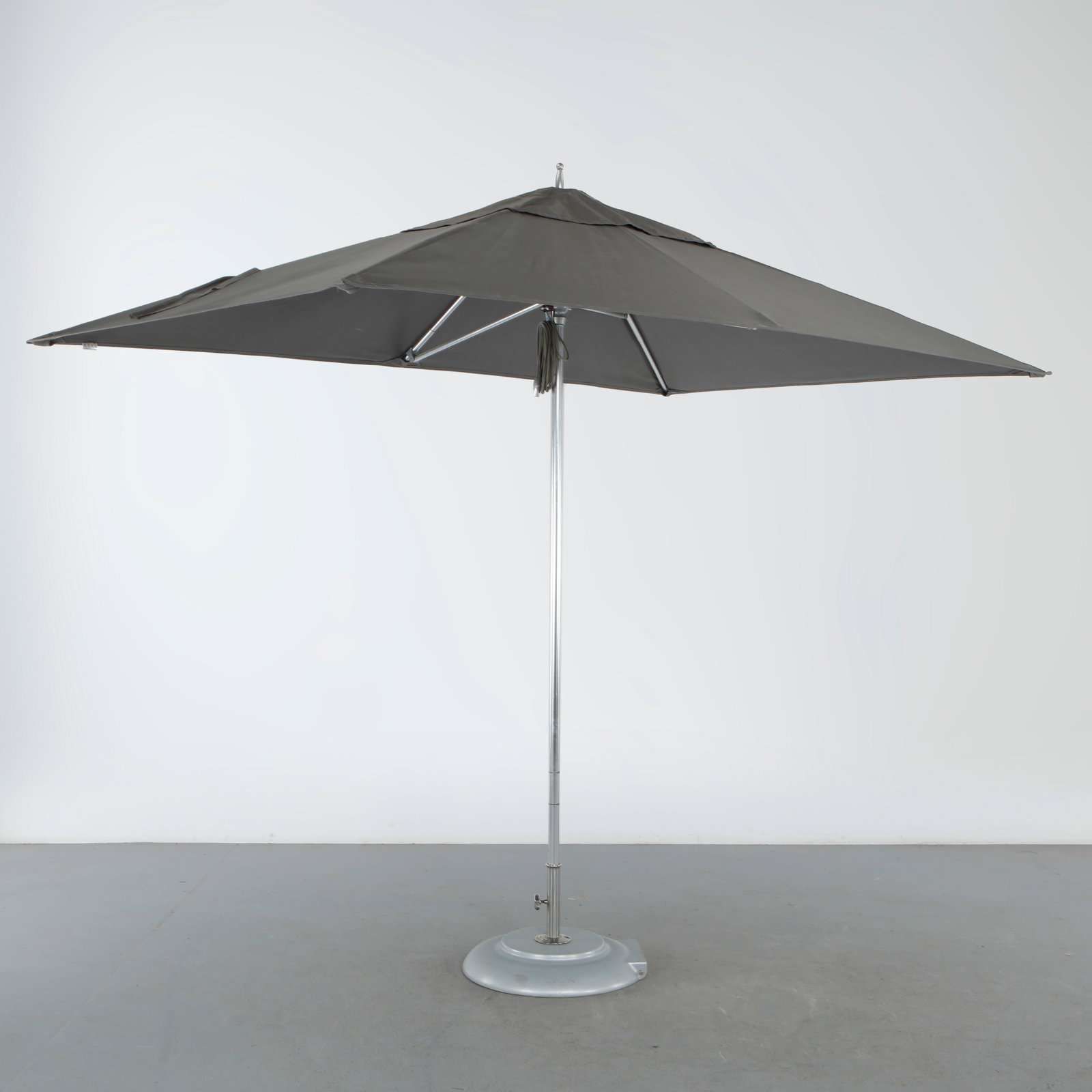 Tuuci Ocean Master Umbrella with Base: A center-pole market umbrella in polished stainless steel with a square gray canopy, opened by a pull-cord mechanism running through the mast, and set on a weighted round base.