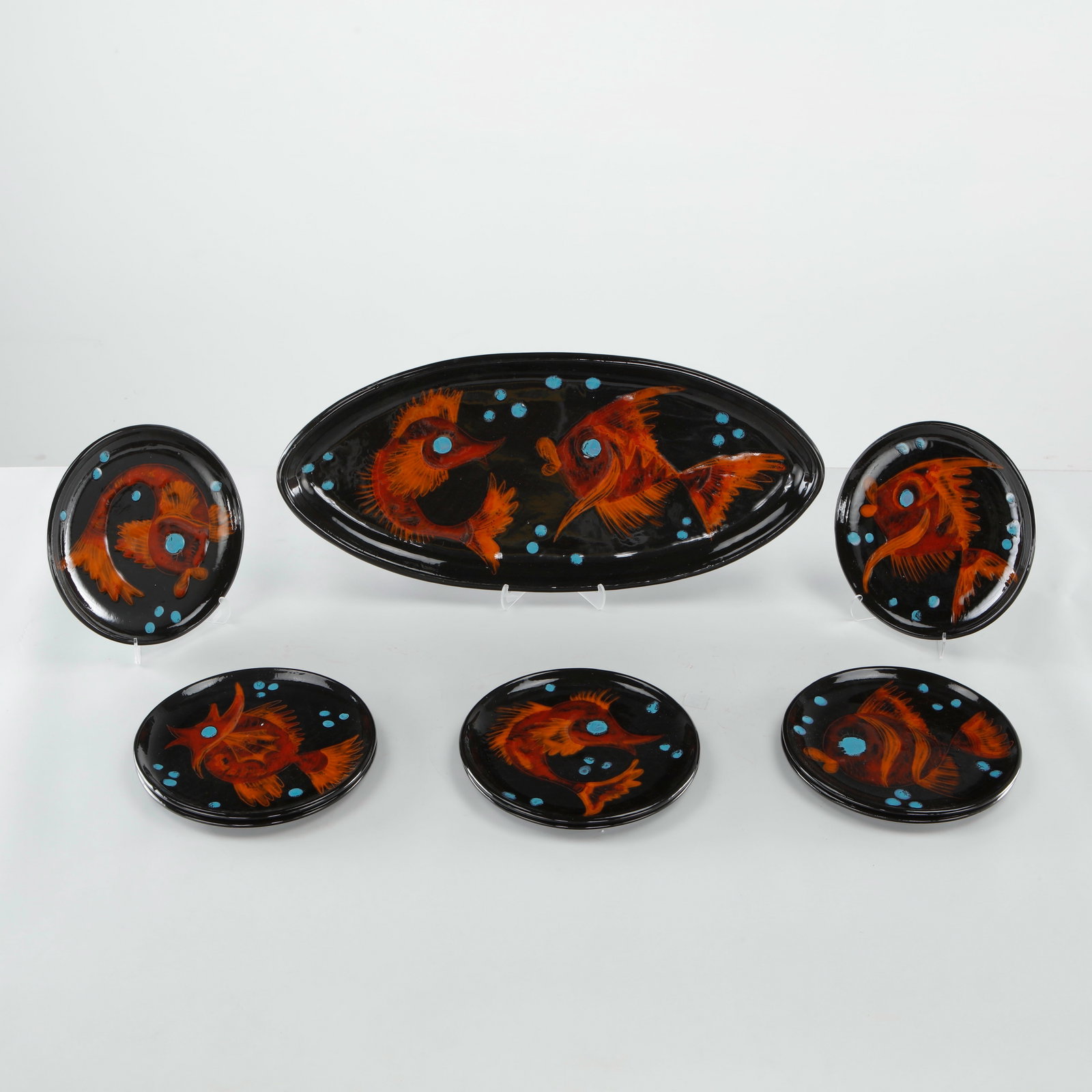 French Ceramic Fish-Motif Dinner Service, Signed, 1960s: A ceramic dinner service comprising one oval platter and eight round plates, each decorated with a hand-painted stylized fish in orange-brown tones against a glossy black ground, accented with raised 