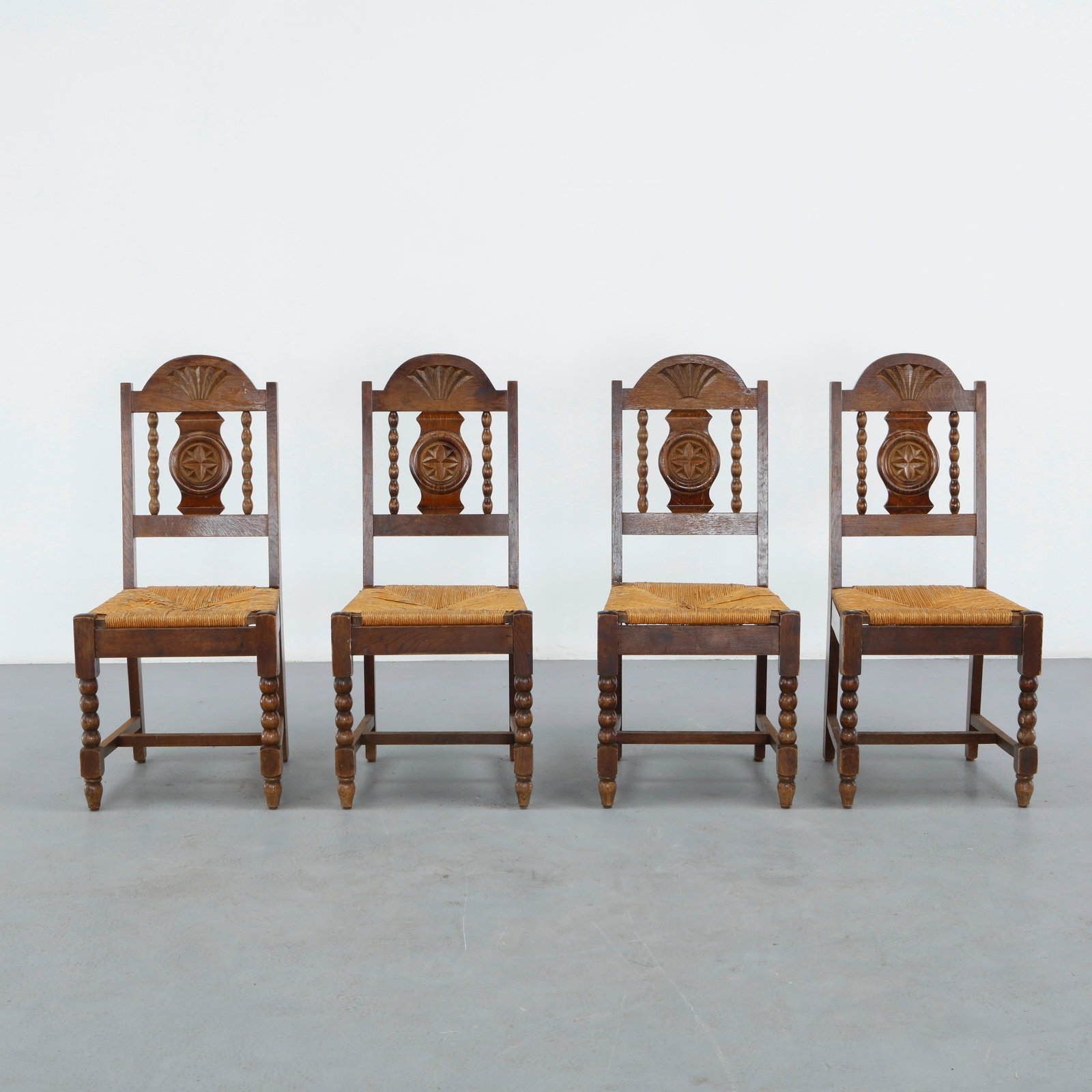 Set of Four Oak Chairs in the style of Charles Dudouyt, 1950s: Four oak chairs in the style of Charles Dudouyt, each with a distinctive carved crest rail, including turned spindle and shell-carved motifs, above a rush seat, forming an eclectic rustic grouping.
