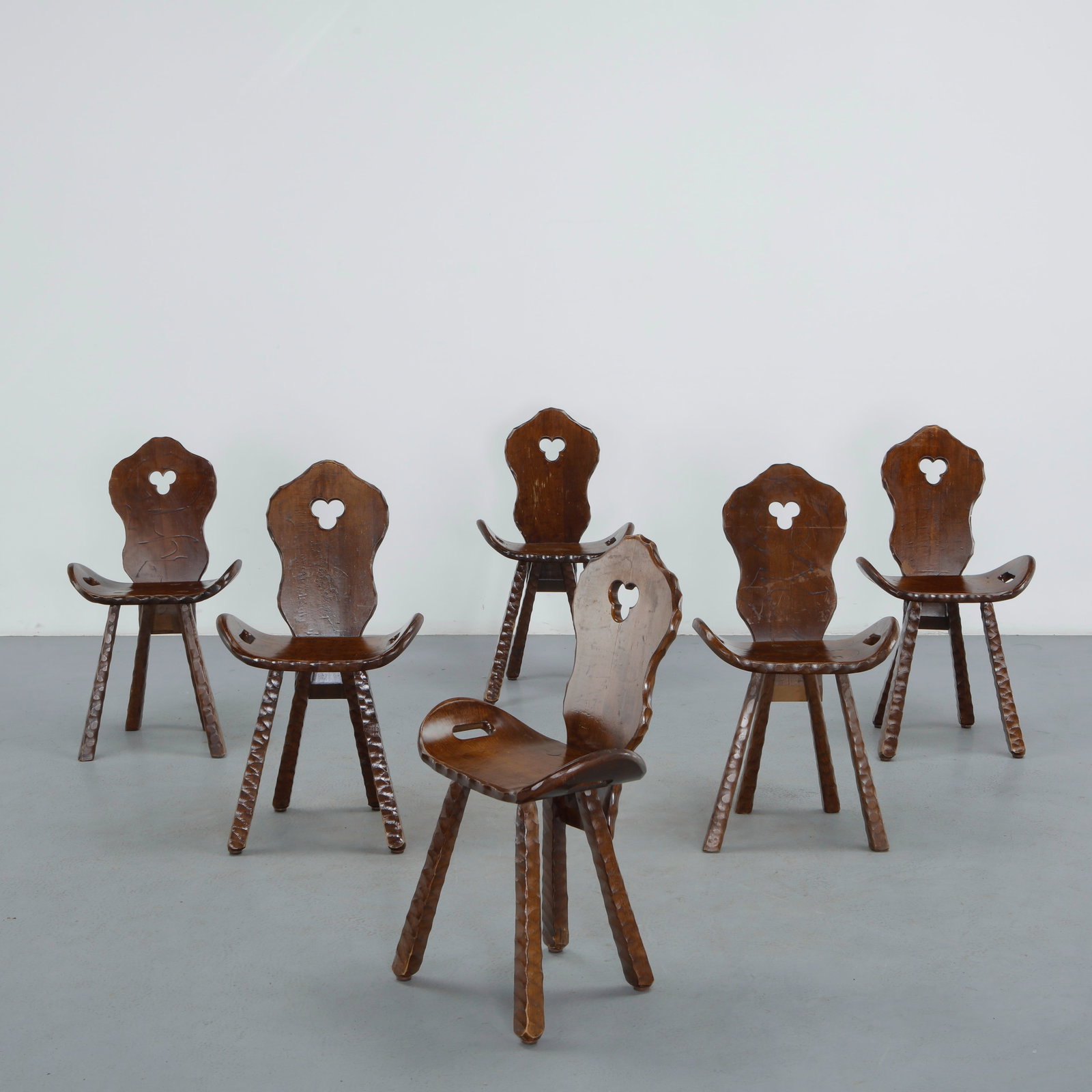Set of Six Brutalist Spanish Chairs, 1960s: Six dark stained Spanish chairs with sculptural, organically carved backrests pierced by a curved cutout motif, resting on tripod style carved legs, a striking, brutalist-inspired rustic design.