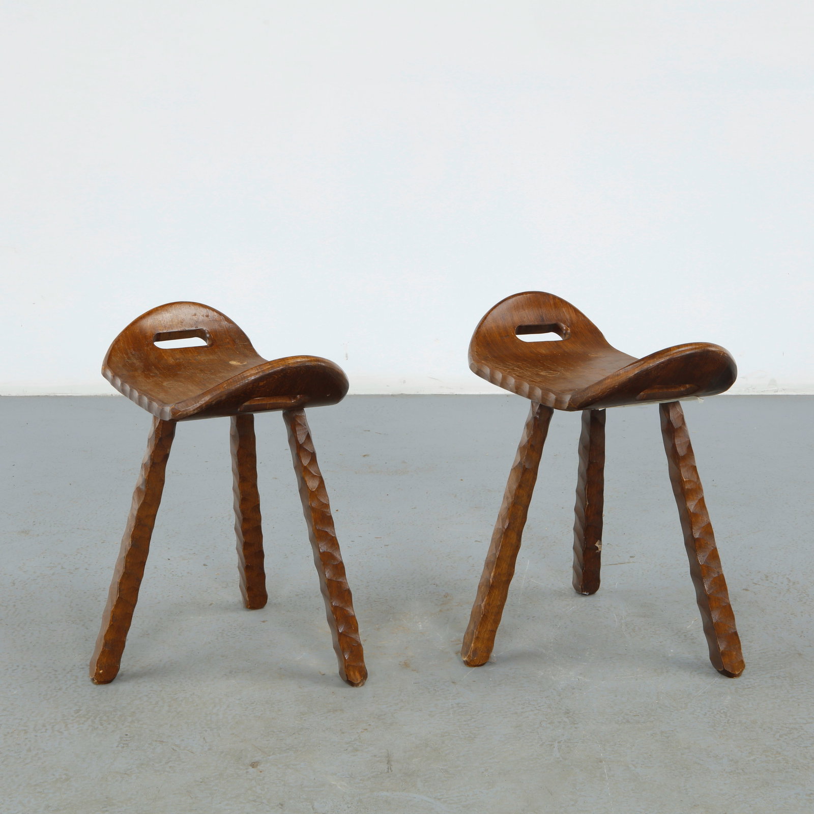 Pair of Brutalist Spanish Stools, 1960s: Hand carved from solid wood, each with a saddle shaped seat pierced by a carrying handle on three faceted legs. Warm wood toned patina and organic, ergonomic profile. Height: 18.25 in. Width: 19.5 in.