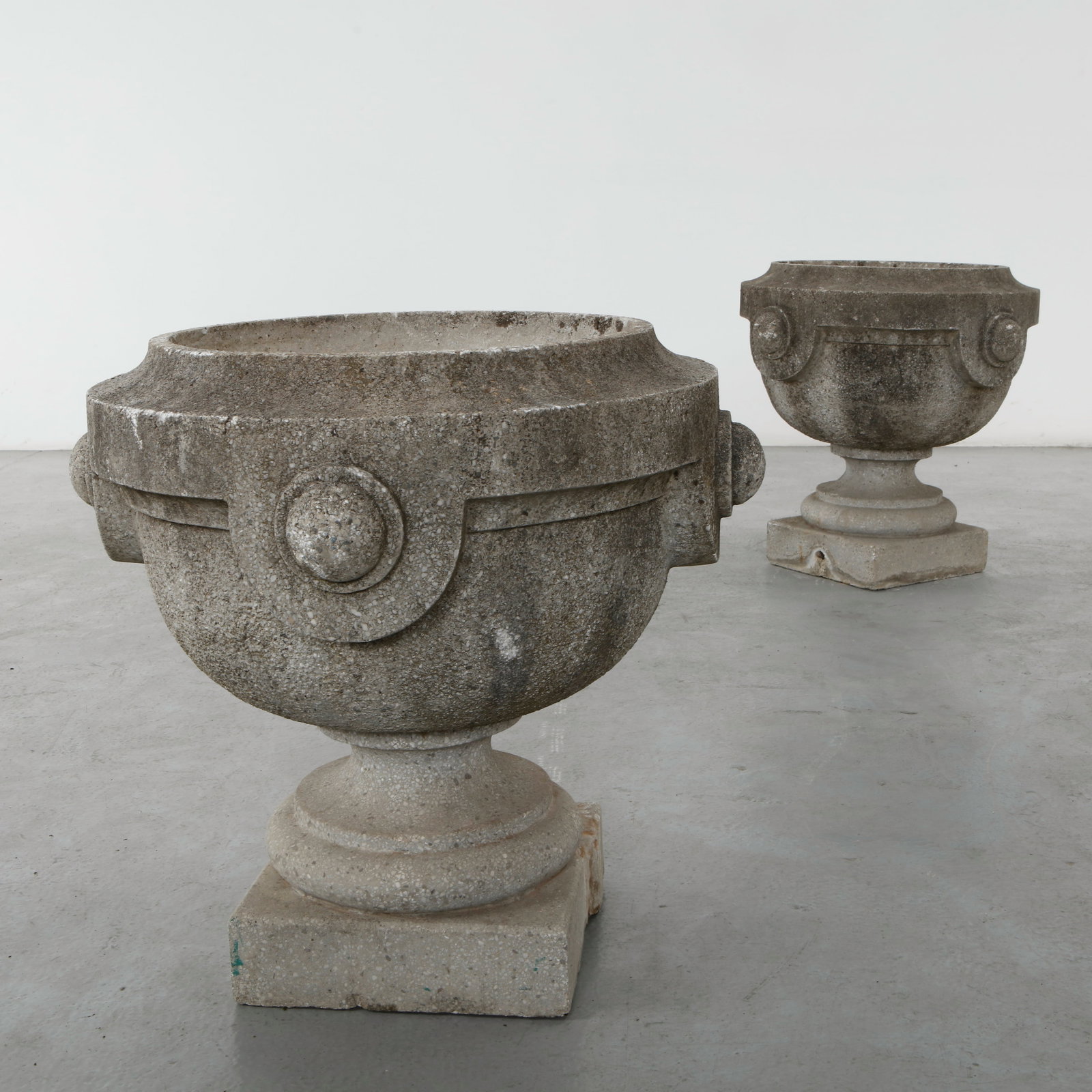 Pair of Monumental Stone Urn Planters, 1960s: Monumental carved stone urn planters with classical scroll handles and pedestal bases. Their restrained neoclassical design is enhanced by a richly aged surface developed through decades of outdoor