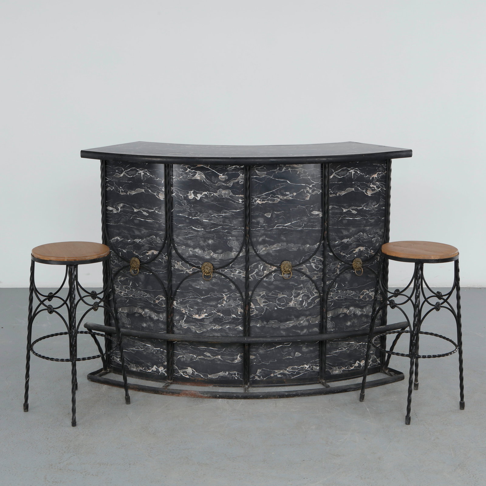 Hollywood Regency Curved Bar with Matching Stools, 1970s: Featuring a sculptural wrought iron frame, this curved bar is finished with black marble-effect panels, brass lion head ring pulls, and a black laminate top. Accompanied by a pair of matching stools, 