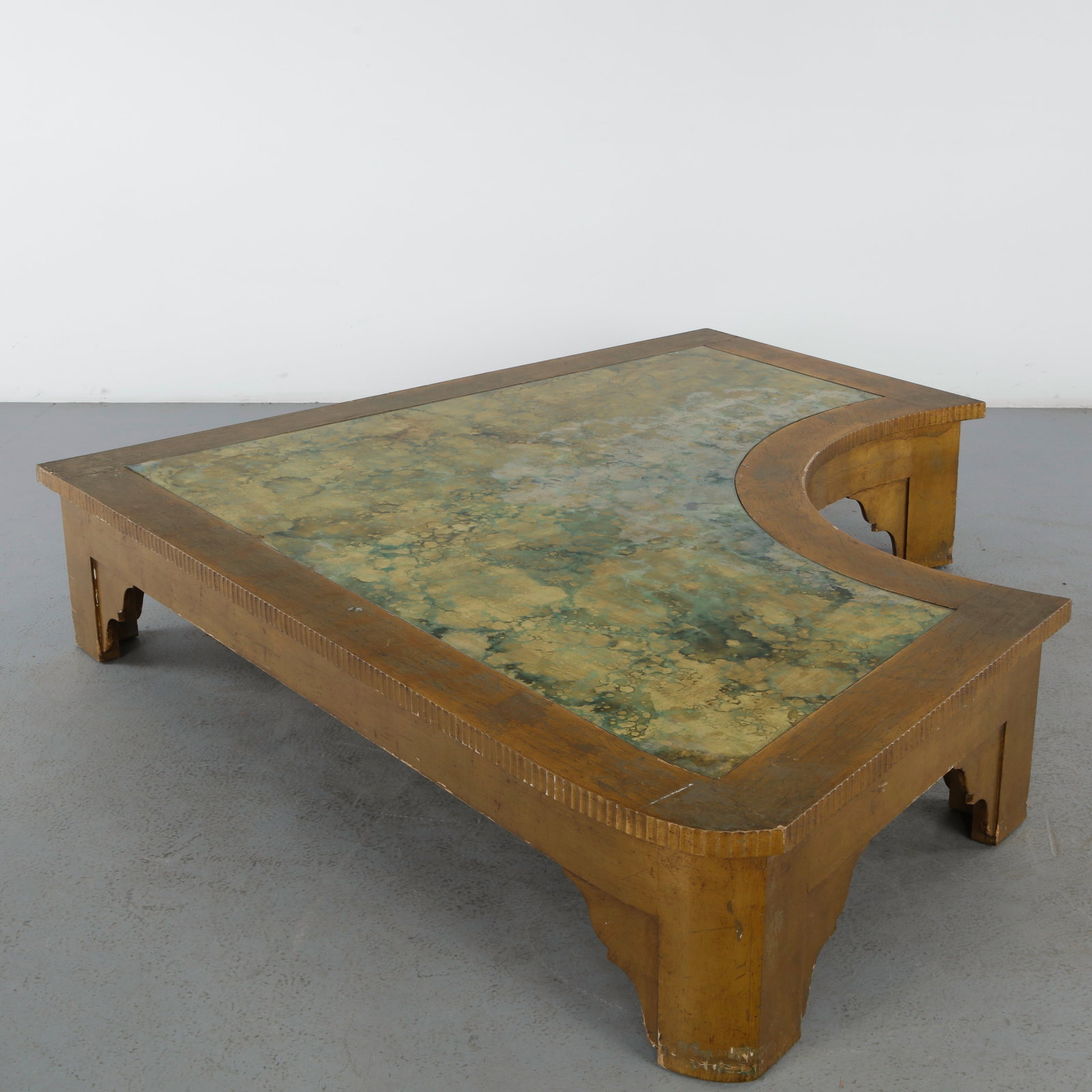 Monumental Hollywood Regency Asymmetrical Coffee Table, 1970s: Monumental asymmetrical coffee table featuring a gilt wood frame and a glass top with a richly textured decorative backing in green and gold hues. The sweeping form and theatrical presence exemplify t