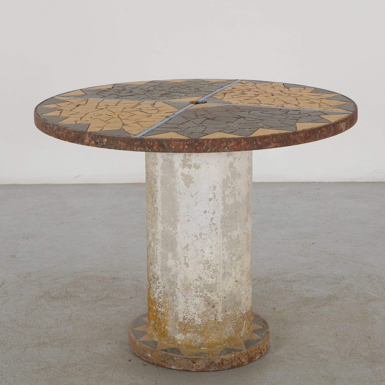 Mosaic Outdoor Dining Table with Geometric Inlay, 1960s: A sculptural outdoor table featuring a circular mosaic top divided into geometric fields of contrasting stone and ceramic fragments. The rusted metal edging and weathered concrete pedestal reinforce i