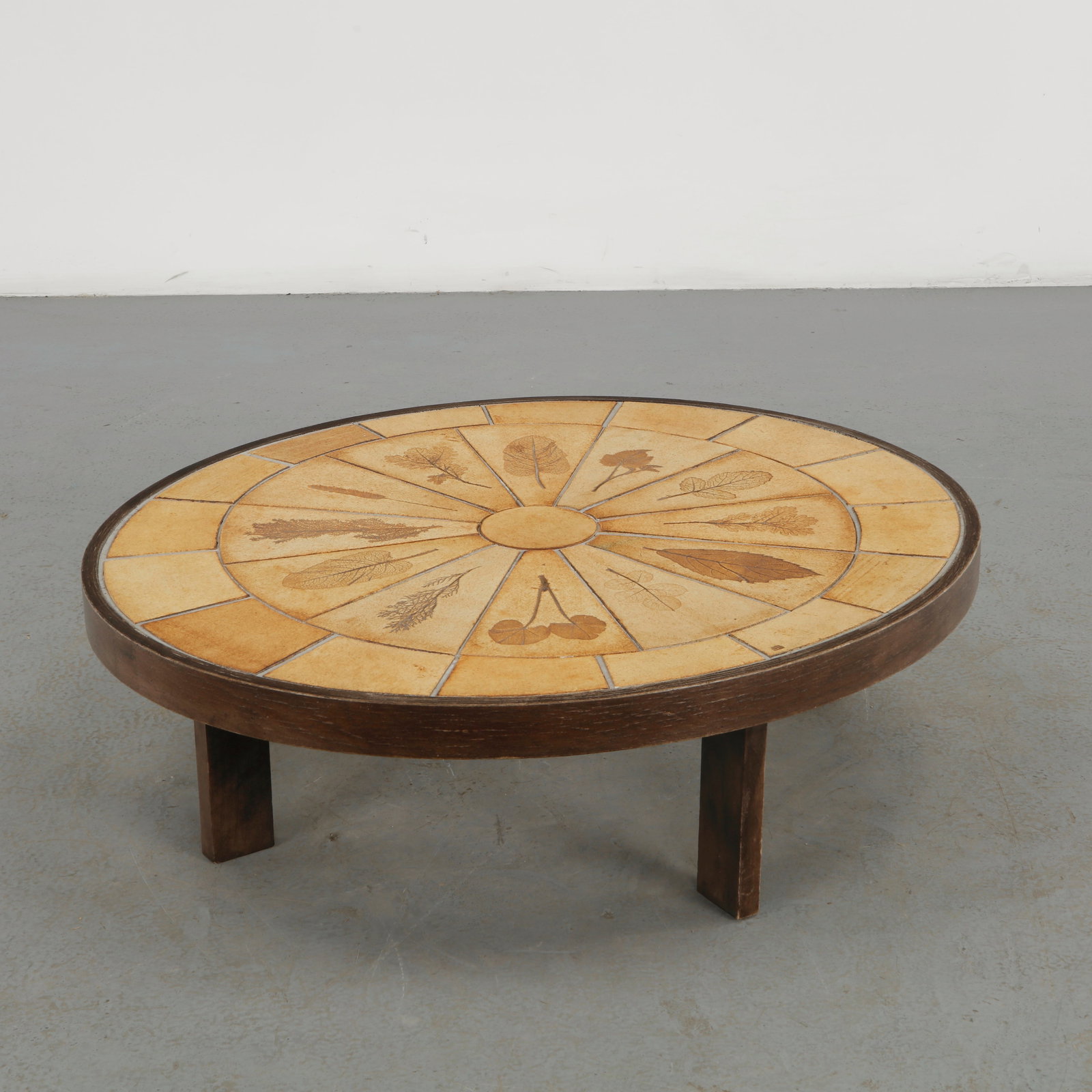 Roger Capron, Ceramic Oval Coffee Table, 1960s: A sculptural oval coffee table featuring a richly decorated ceramic top composed of radiating geometric segments and botanical motifs, set within a dark wooden frame. The hand rendered surface combine