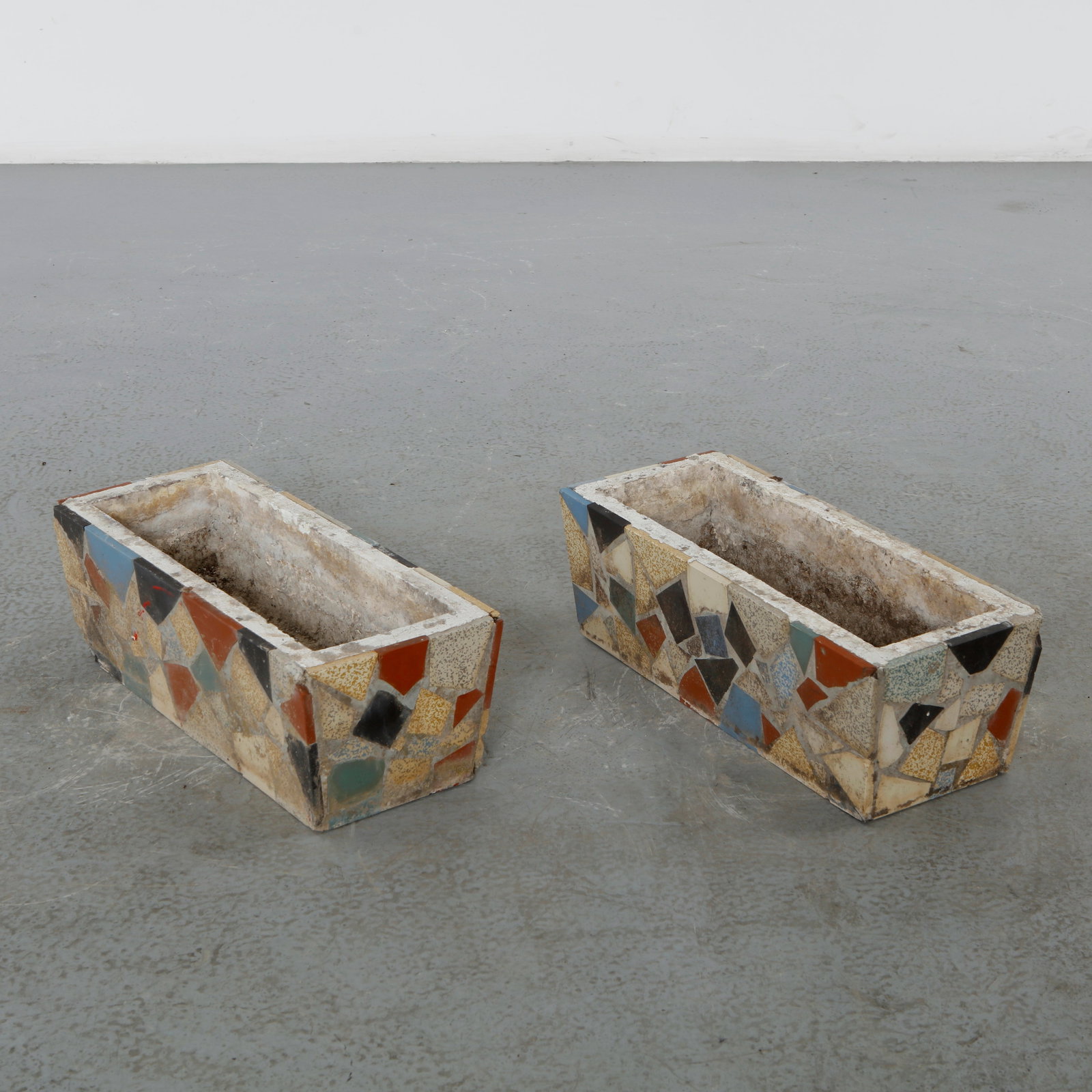 Pair of Mosaic Tile Planters, 1960s: Rectangular concrete planters clad in a hand-set mosaic of broken glazed tile fragments in cream, ochre, blue, and black. Each bears a distinct pattern and a rich, weathered patina from decades outdoo