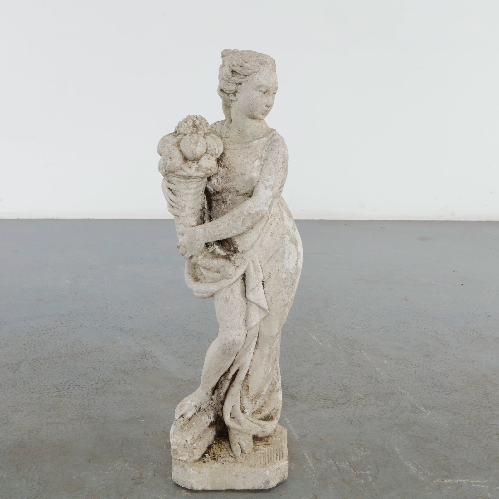 Classical Garden Statue of a Maiden with Cornucopia, 20th Century: A graceful cast stone garden figure depicting a classical maiden holding a cornucopia overflowing with fruit, symbolizing abundance and prosperity. The naturally weathered surface lends the sculpture 
