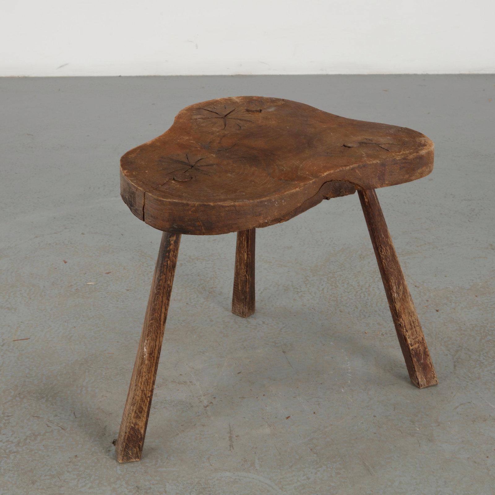 Brutalist Live Edge Wood Stool, 1960s: A singular three-legged stool carved from a substantial irregular timber slab, with pronounced natural fissures and a richly weathered surface. The uncompromising use of raw wood gives the piece a