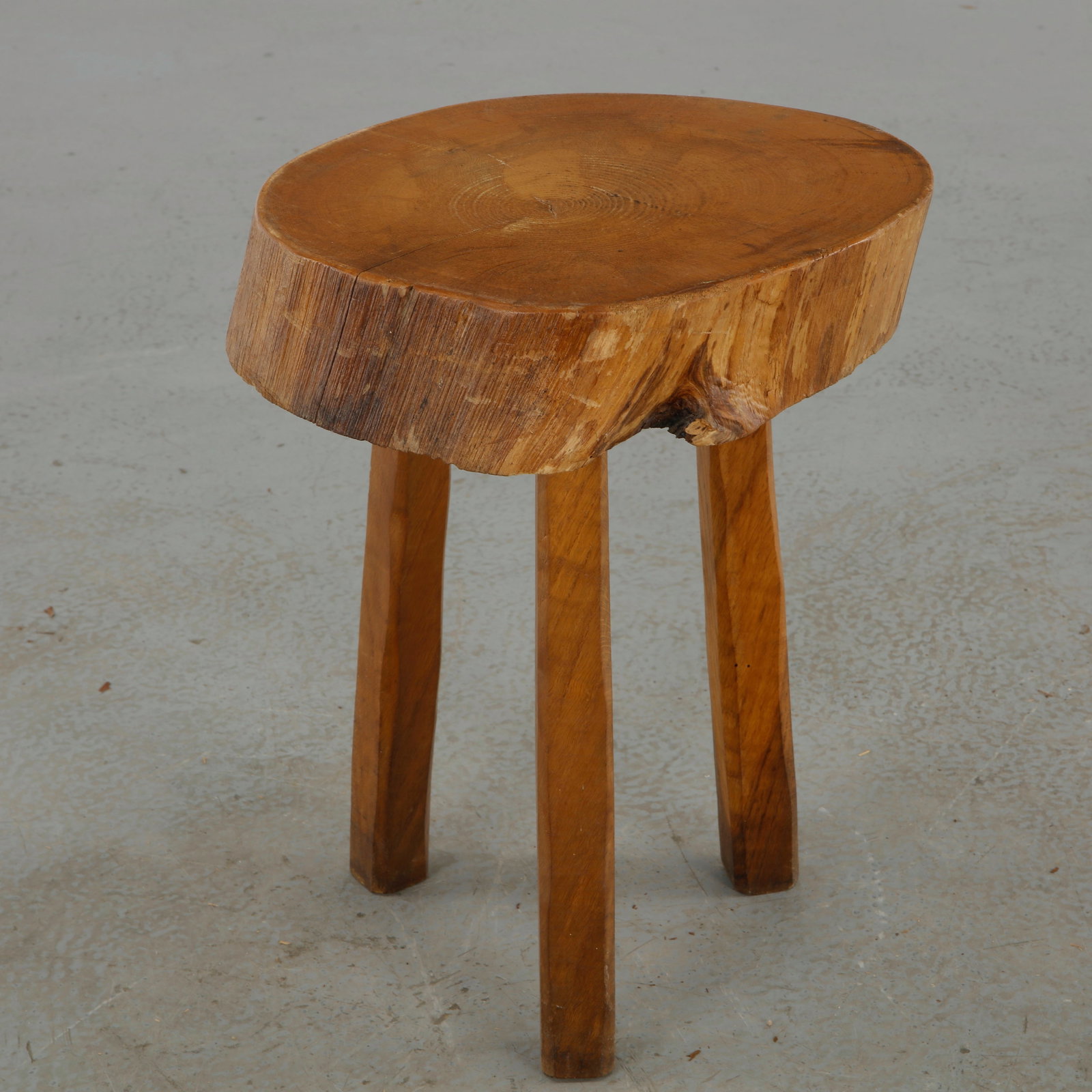 Primitive Wooden Stool, 1960s: A substantial primitive stool fashioned from a thick section of timber, with a naturally irregular edge and strongly pronounced grain. Its straightforward three-legged construction preserves the