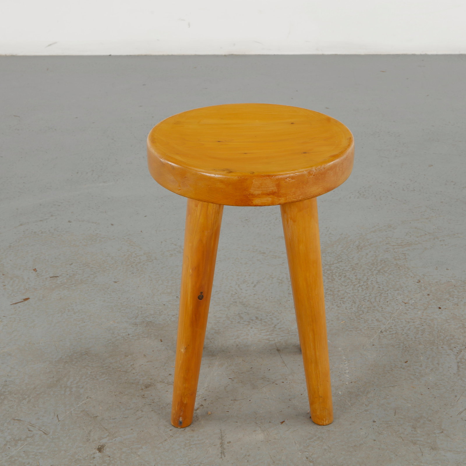 Primitive Wooden Stool, 1960s: A restrained rustic stool crafted from solid wood, distinguished by a thick rounded seat and gently splayed legs. The warm honey-colored patina and visible grain emphasize the tactile quality and unde