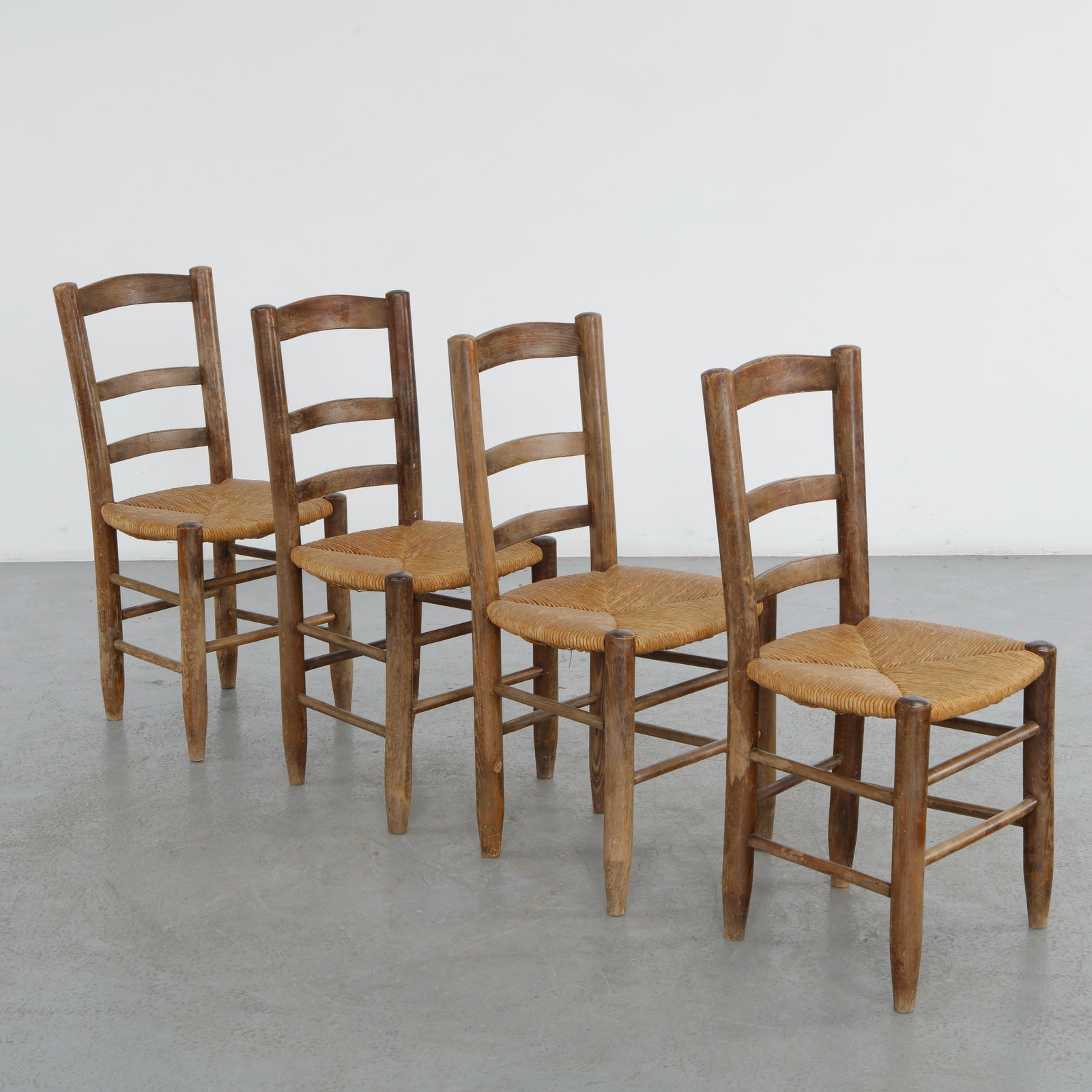 Set of Four Chairs in the style of Charlotte Perriand, 1960s: Crafted in solid wood with hand-woven rush seats, these ladder-back chairs embody the restrained elegance of French mountain modernism. Their honest construction and warm patina evoke the timeless