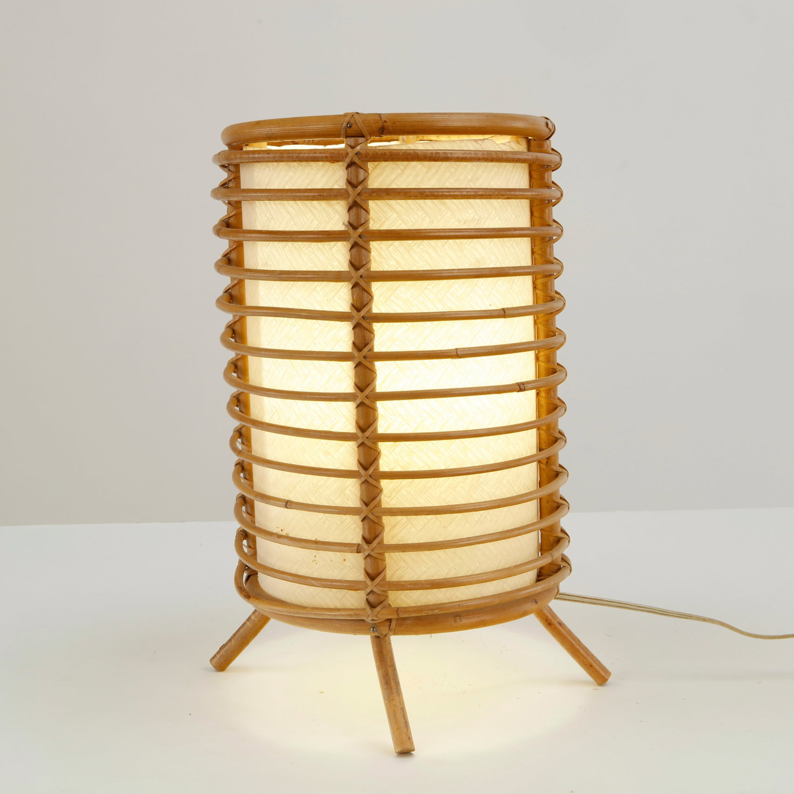 Louis Sognot, Rattan Cylindrical Table Lamp, 1960s: Executed in bamboo and woven rattan, this table lamp features a cylindrical diffuser enclosed within a sculptural cage of bent bamboo rings. The rhythmic structure rests on a delicate tripod base, hig