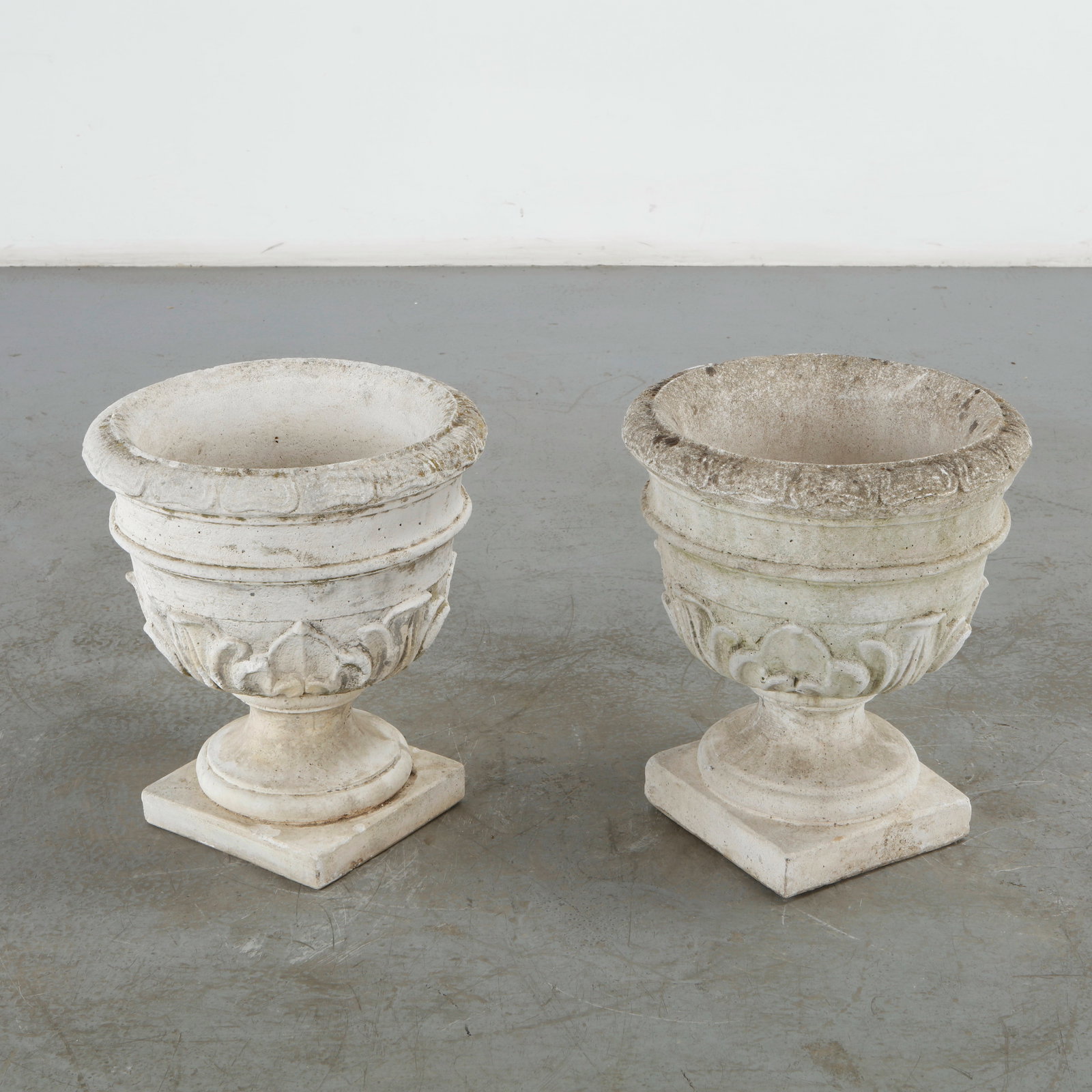 Pair of Stone Urn Planters, 1960s: This pair of cast stone urn planters showcases a beautiful Neoclassical style, highly valued for mid-century garden ornamentation. It is intricately molded with a bold relief pattern of acanthus