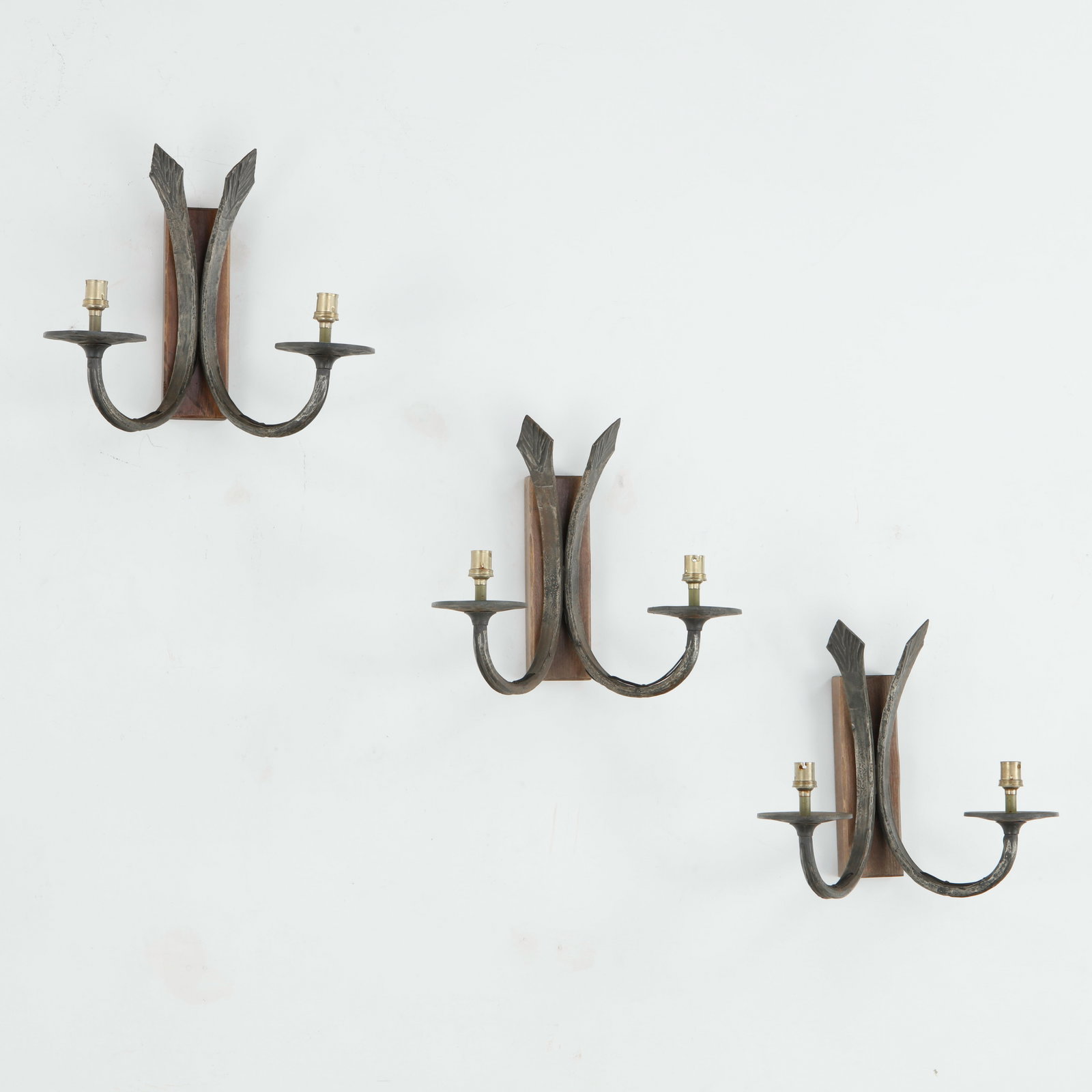 Set of Three Brutalist Bronze Double-Arm Wall Sconces: Set of three sculptural double-arm wall sconces in bronze, each mounted on a rectangular wooden backplate. The sweeping curved arms and elongated, textured vertical elements create a striking Brutalis