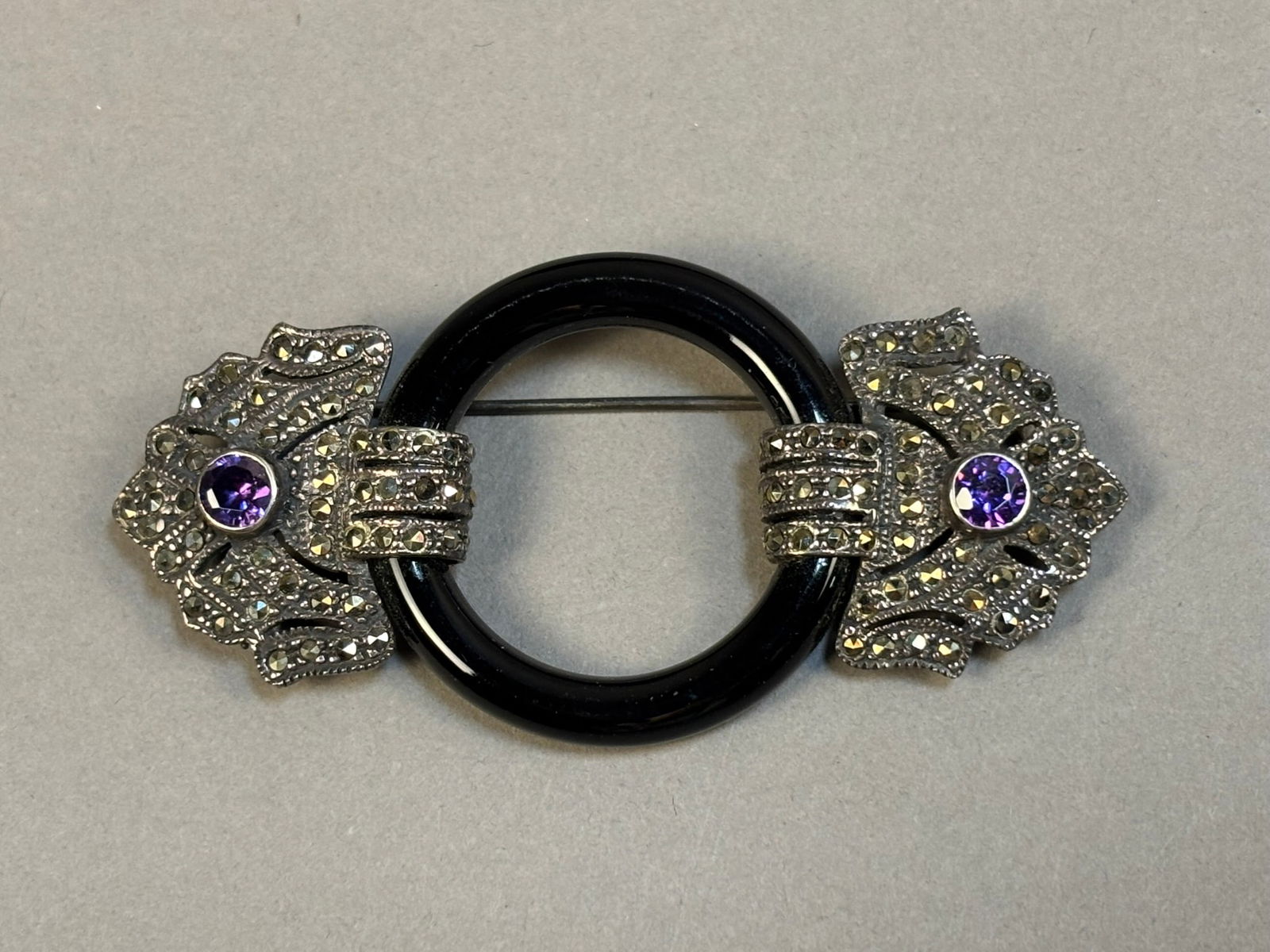 Art Deco Sterling Marcasite Amethyst Black Glass Circle Pendant Brooch: Art Deco style sterling silver brooch featuring ornate marcasite set floral terminals each centered with round faceted purple stones consistent with amethyst. The decorative ends are joined by a polis