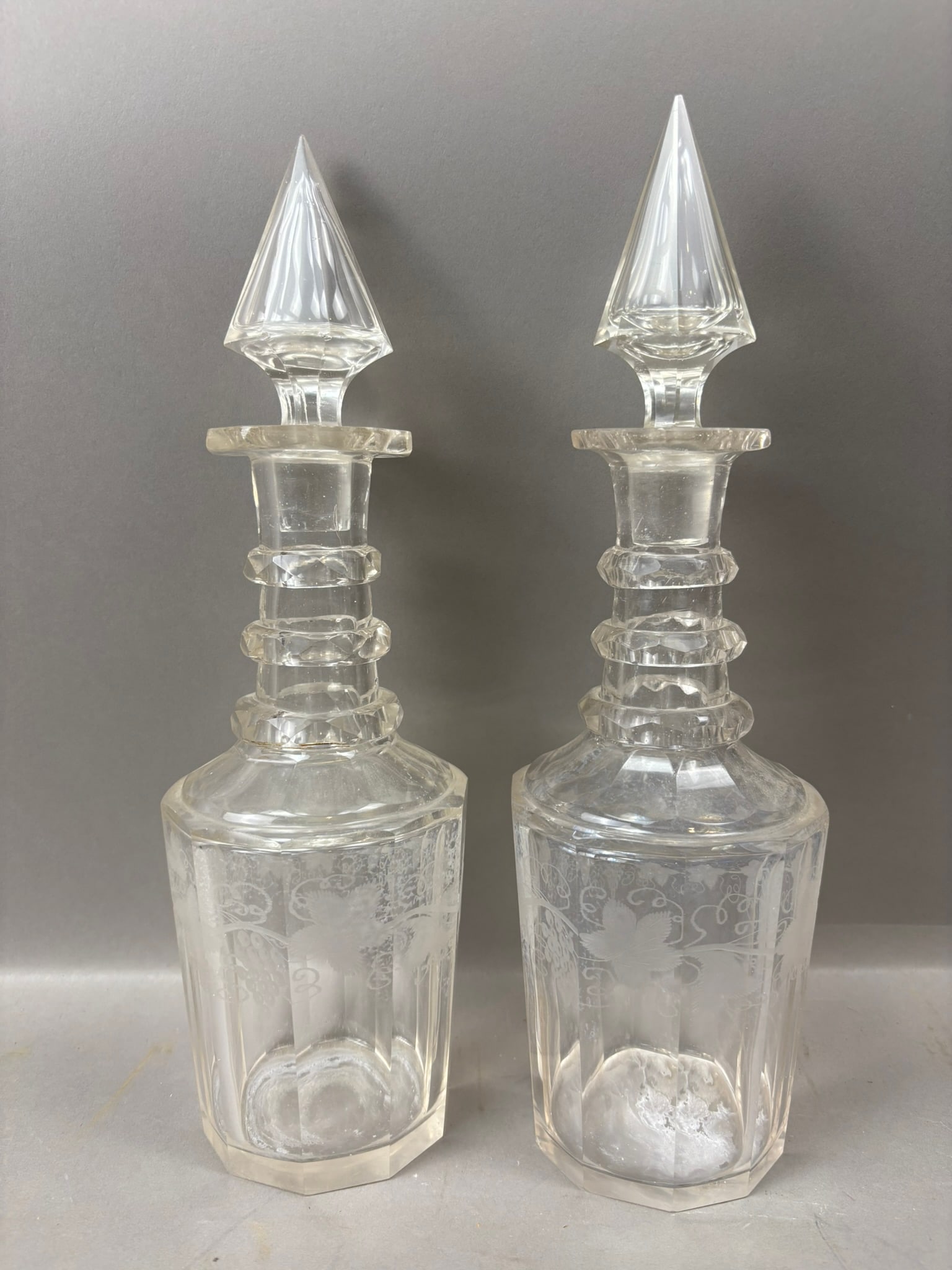 Pair Antique Etched Glass Decanter Bottles w/ Faceted Stoppers 12": Pair of antique clear glass decanter bottles featuring paneled bodies with delicate etched floral and scrolling decoration, each fitted with tall faceted glass stoppers of pointed form. The bottles me