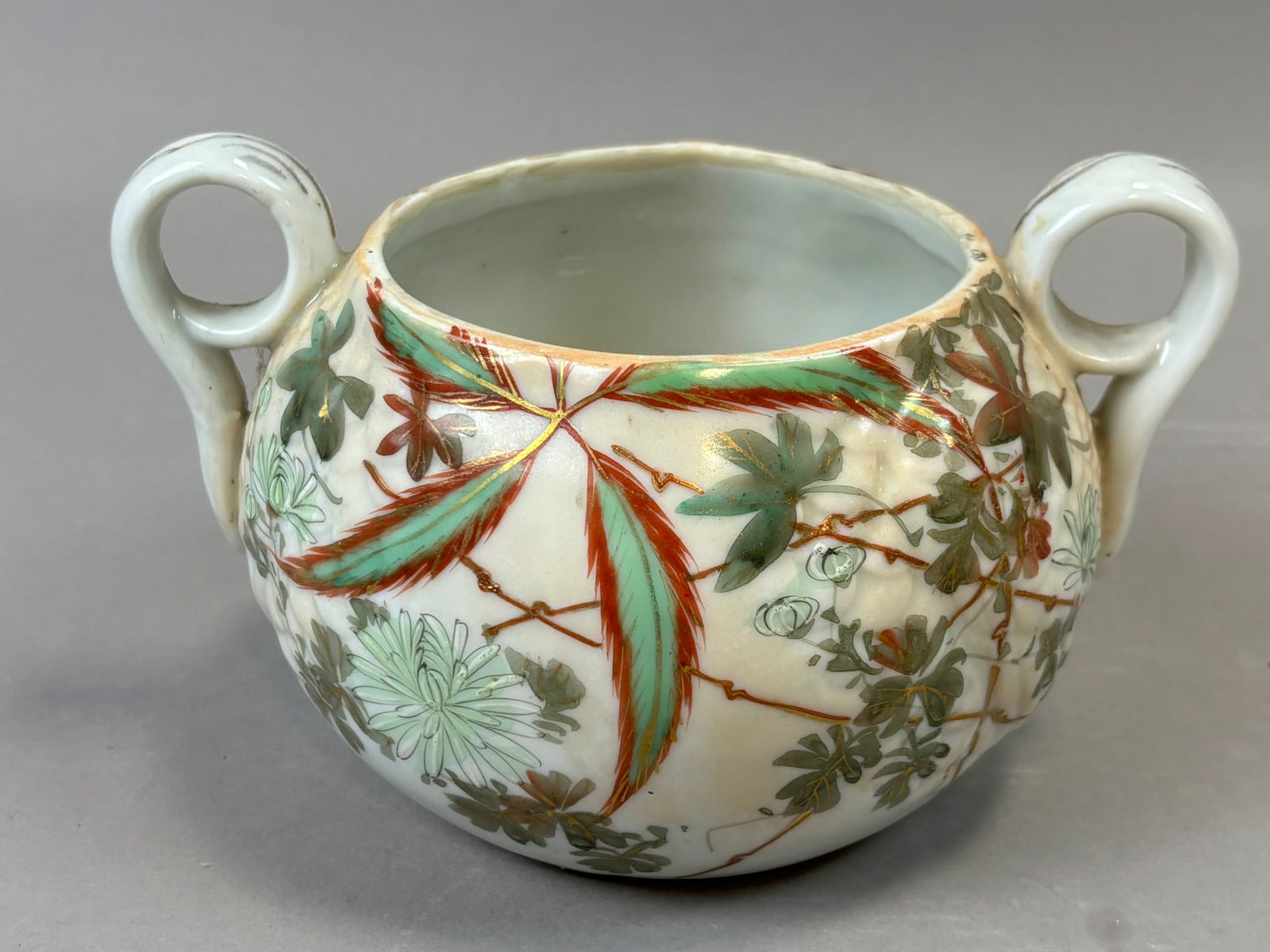 Japanese Satsuma Style Two Handle Enamel Decorated Jar Meiji Period: Japanese Satsuma style porcelain jar with twin loop handles and relief molded body decorated in overglaze enamels and gilt featuring chrysanthemum blossoms, maple leaves, and trailing foliage. The rou