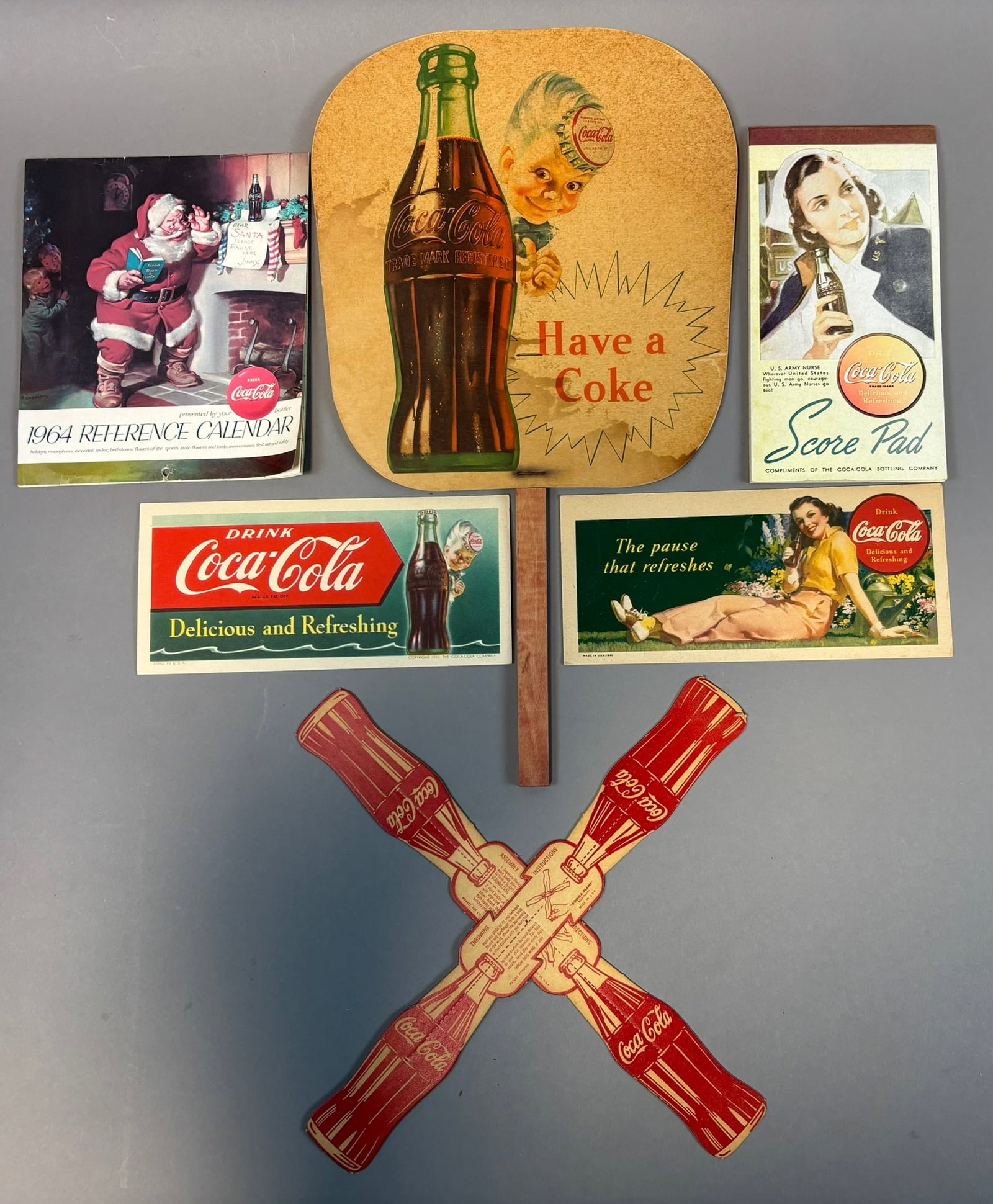 Lot 6 Vintage Coca Cola Advertising Items Score Pad Blotters Fan & Spinner: Assorted vintage Coca-Cola paper advertising lot including a die-cut ?Have a Coke? hand fan with wooden handle, a 1964 Coca-Cola reference calendar featuring Santa Claus imagery, a Coca-Cola score pad