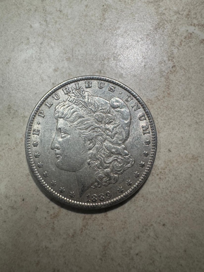 1889 Morgan Dollar AU50 (Non-Graded): The 1889 Morgan Silver Dollar in AU50 condition is a lightly circulated coin that still retains much of its original mint luster, especially around protected areas like the hair above Liberty?s ear an