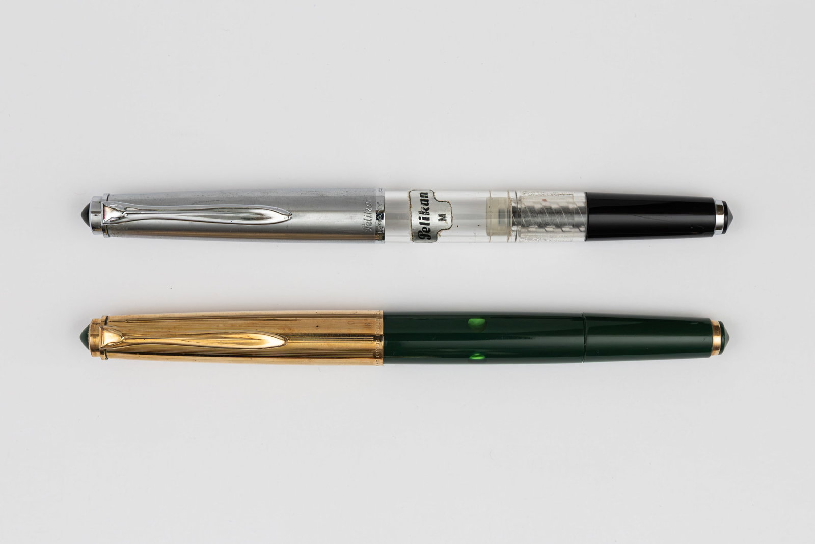 Pelikan P1 Gold Cap & Demonstrator Fountain Pens: Pelikan P1 fountain pens, circa 1960’s. Pelikan P1 fountain pens in green with gold filled cap and demonstrator models, both with 14K gold nibs. Length 5 1/4” and 5 3/4” posted, filling 