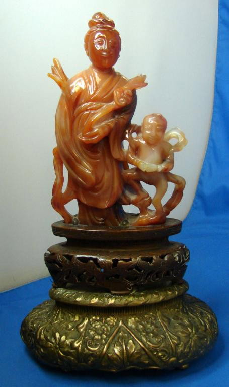 Carved Carnelian Kwan Yin w/Small Boy: Carved Carnelian figurine of Kwan Yin with a small boy at her side, sits on wooden base which sits on an ornate bronze base. 10 1/2" tall, including base; figure is 6" tall.
