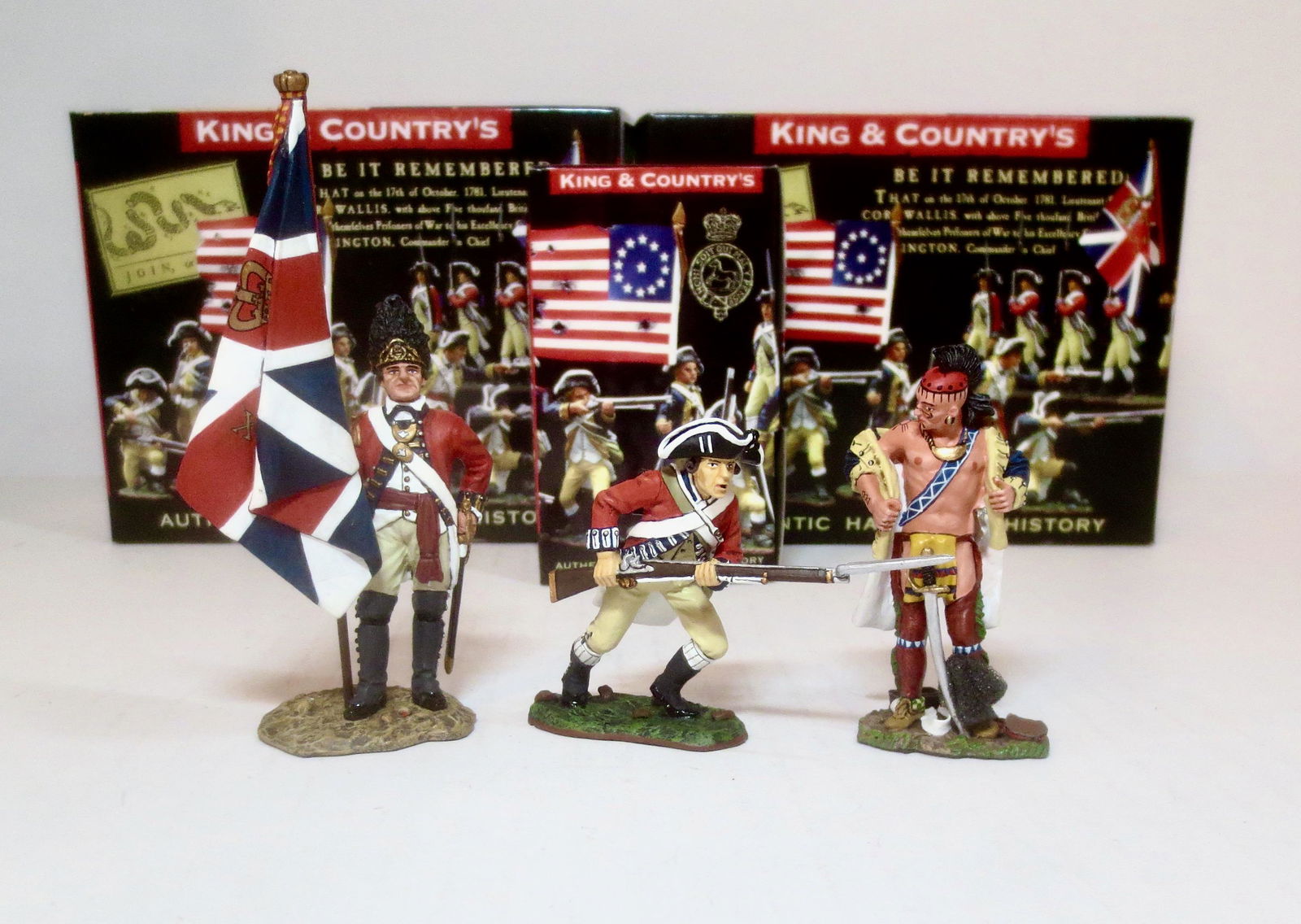 King & Country American Revolution Series: RWF Officer with Regimental Colour #BR082, British Infantry Running with Rifle #BR049, and Woodland Indian trying on a Continental Jacket #BR052. 3 pieces. Flag flaking paint, otherwise all in mint co