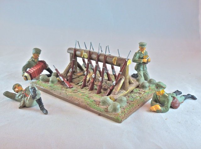 Rifle/Helmet Stand+4 Elastolin Camp Figures: Postwar stand with 12 rifles and helmets is about 10 years old. Each rifle is removable and cast metal. The soldiers on back, playing concertina and kneeling cutting bread are all Lineol and have hard