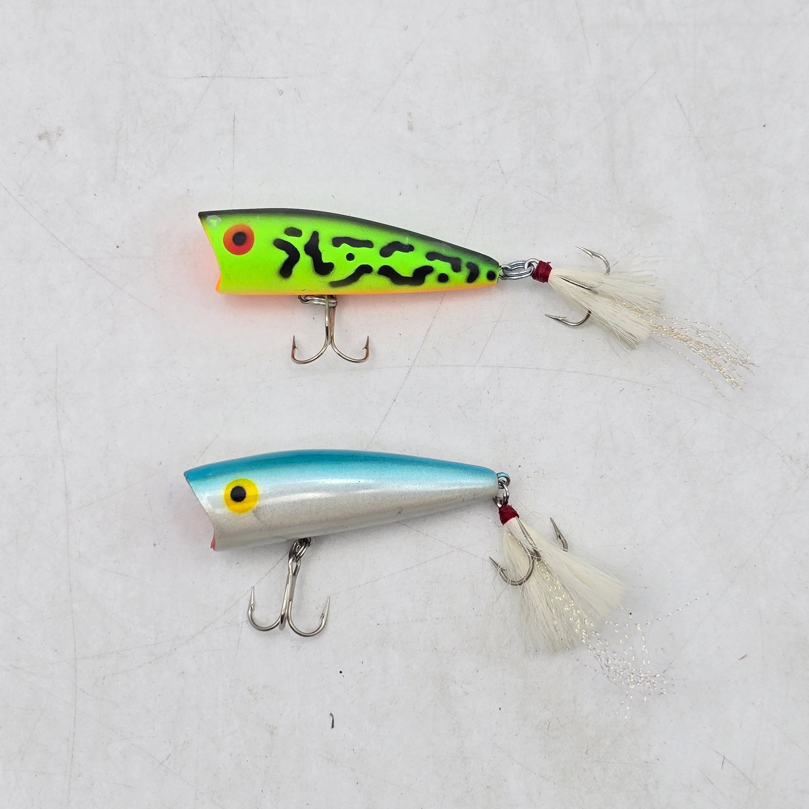 Topwater Fishing Lures 2pc: Lot of 2 Topwater Fishing Lures. Includes popper-style lures in bright frog pattern and classic silver finish, both featuring feathered treble hooks. Designed for surface action and aggressive