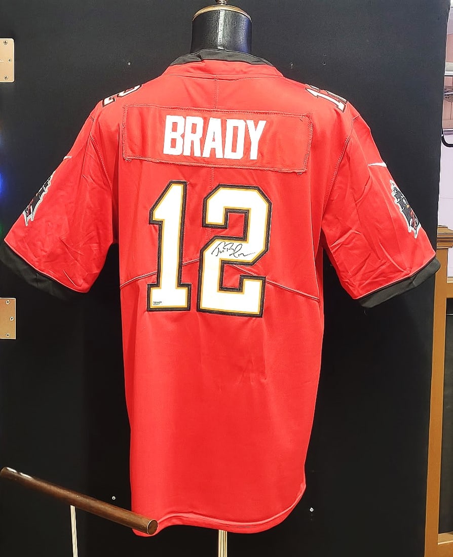 TOM BRADY SIGNED AUTHENTICATED TAMPA BAY JERSEY: The image shows a red Tampa Bay Buccaneers jersey with the number 12, which was Brady's number during his time with the team. The signature is consistent with his known autograph, a flowing cursive si