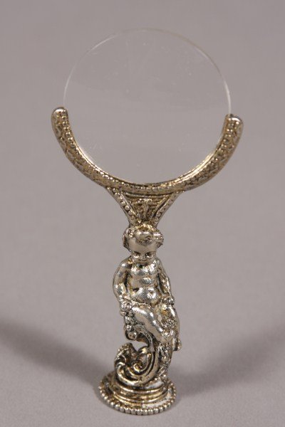 Molded White Metal Magnifying Glass, 20th C. In the : In the form of a putti supporting a magnifying lens. Height: 5 1/2" Condition: Minor scratches to lens.